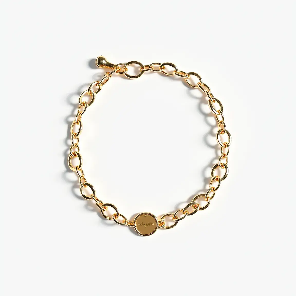 This ankle bracelet features a series of interlocking gold-toned metal links, creating a seamless chain. At the center, it has a small, round, flat charm also made of the same gold-toned material. The charm appears to be inscribed or engraved, adding a personalized touch. The ankle bracelet is fastened with a toggle clasp, where a small bar is threaded through a loop to secure it in place. The simplicity and elegance of the design make it a versatile accessory for various occasions.