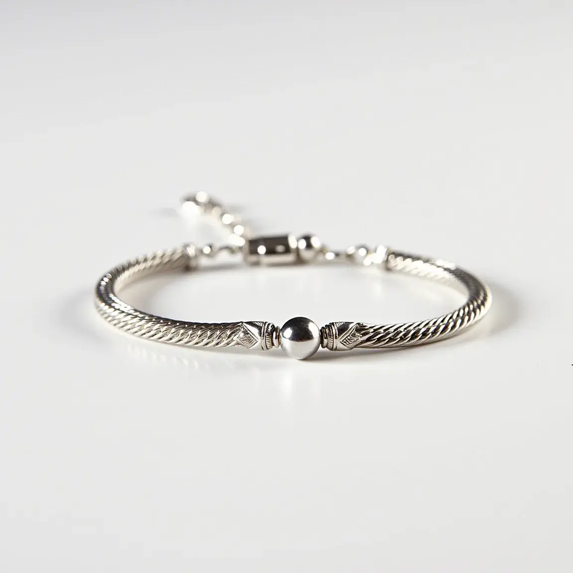 This ankle bracelet features an intricately braided or twisted design, primarily composed of a metallic material, possibly silver or white gold, given its sheen and color. At the center, it is adorned with a smooth spherical bead, which serves as a focal point of the bracelet's design. The ends of the braided section transition into polished caps, incorporating detailed engravings for added elegance. The bracelet is secured with a lobster claw clasp, providing both security and ease of use. The overall craftsmanship suggests a combination of both modern and traditional design elements, making it a versatile accessory.