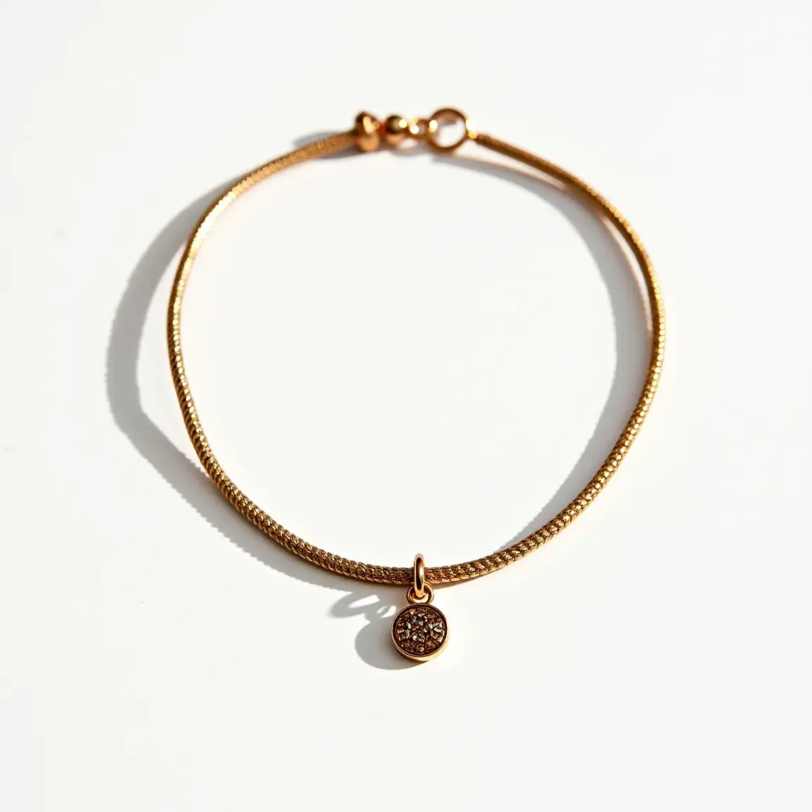 This ankle bracelet features a delicate, finely woven gold-colored chain and is adorned with a circular charm. The charm is encrusted with what appear to be small, round-cut stones set closely together, suggesting a pave setting which provides a sparkling effect. The bracelet is secured with a simple, circular clasp which adds to its overall elegant and minimalist design.