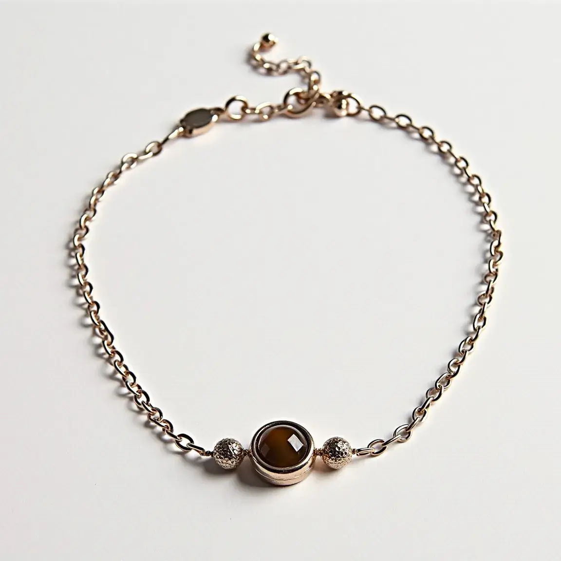 This ankle bracelet features a delicate chain crafted from a metal material, possibly gold or gold-plated. The centerpiece is a round, bezel-set gemstone that appears to be brown in color, flanked by two textured metal beads. The clasp is adjustable, ending in a small chain with a single bead, allowing for a customized fit. This design showcases a balance of simplicity and elegance, making it a versatile piece for various occasions.