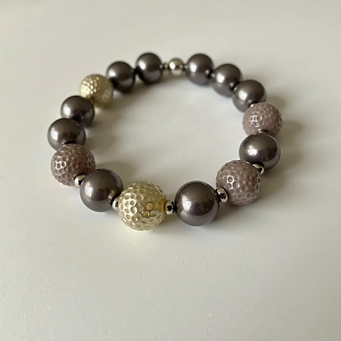 This baby bracelet features a series of spherical beads in various finishes and textures. The beads alternate between smooth, metallic gray spheres and textured, dimpled beads in hues of pearly white and light pink. The bracelet is likely threaded on a stretchable elastic, as there is no visible clasp or attachment mechanism, ensuring ease of wear for a baby. Small gray metallic spacer beads are positioned between each of the larger beads. The combination of smooth and dimpled beads offers an interesting contrast, making the bracelet visually appealing and suitable for a baby.