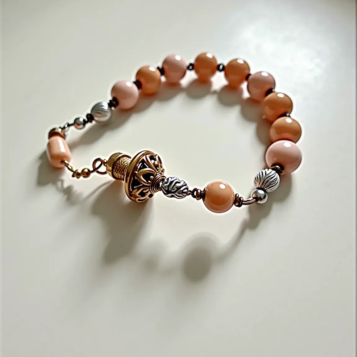 This baby bracelet features a series of smooth, spherical beads in a soft, peachy hue, likely crafted from polished stones or synthetic materials. Interspersed at intervals are metal beads with a textured, ribbed design, accentuating the overall visual appeal. The bracelet is anchored with a prominent, ornate clasp that seems to be a screw-on or threaded type, offering both security and an elegant finish. The detailed design on the clasp and the adjoining components suggest intricate metalwork, adding a touch of sophistication to the otherwise simple yet charming accessory.