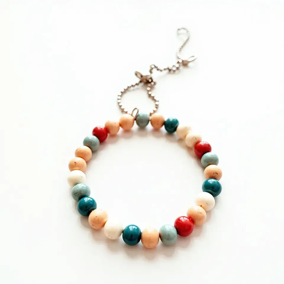 This baby bracelet features a series of round beads strung together in alternating colors of natural beige, vibrant red, soft turquoise, and deep teal. The beads are smooth and uniform in shape, likely made from a mix of wooden and plastic materials. The bracelet is secured with a delicate silver-colored clasp and a short linked chain, allowing for an adjustable fit. The simple yet charming design makes it a cute and versatile accessory for a baby.