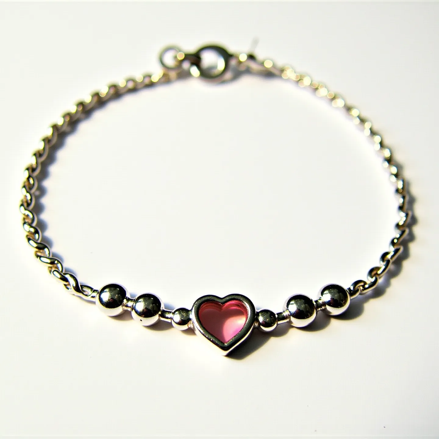 This baby bracelet is crafted from a shiny, silver-toned metal chain featuring a series of small and large round beads. At the center of the bracelet is a heart-shaped charm, which contains a pink gemstone set within a silver frame. The gemstone is smooth and polished with a simple, yet elegant cut that enhances its delicate appearance. The bracelet is secured with a round spring clasp, making it easy to put on and take off.