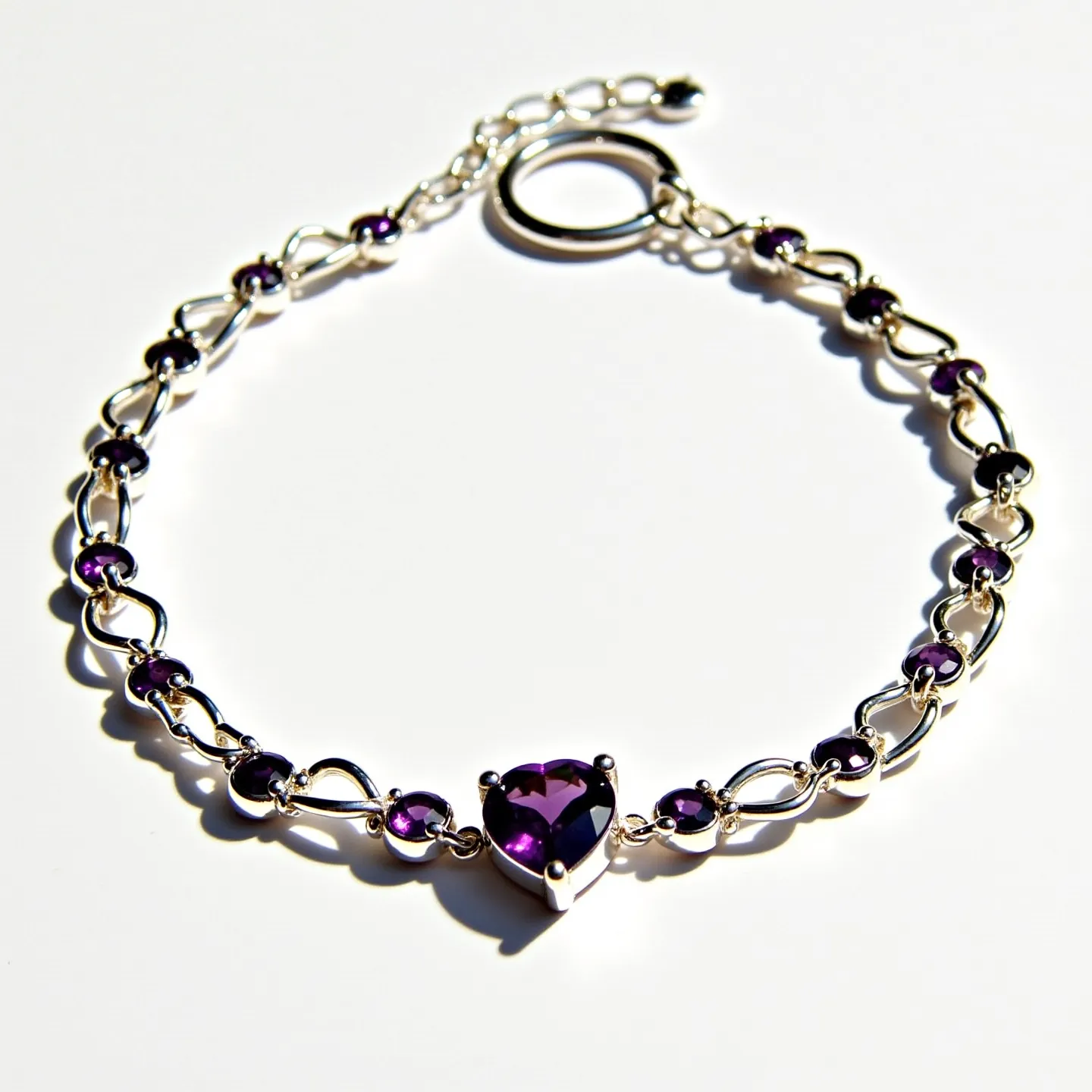 This baby bracelet features a delicate chain crafted from silver. It is adorned with small, round-cut purple gemstones that are evenly spaced along its length. At the center of the bracelet is a heart-shaped purple gemstone set in a three-prong setting, adding a charming focal point to the design. The bracelet is secured with a circular clasp that allows for easy adjustment of its size.