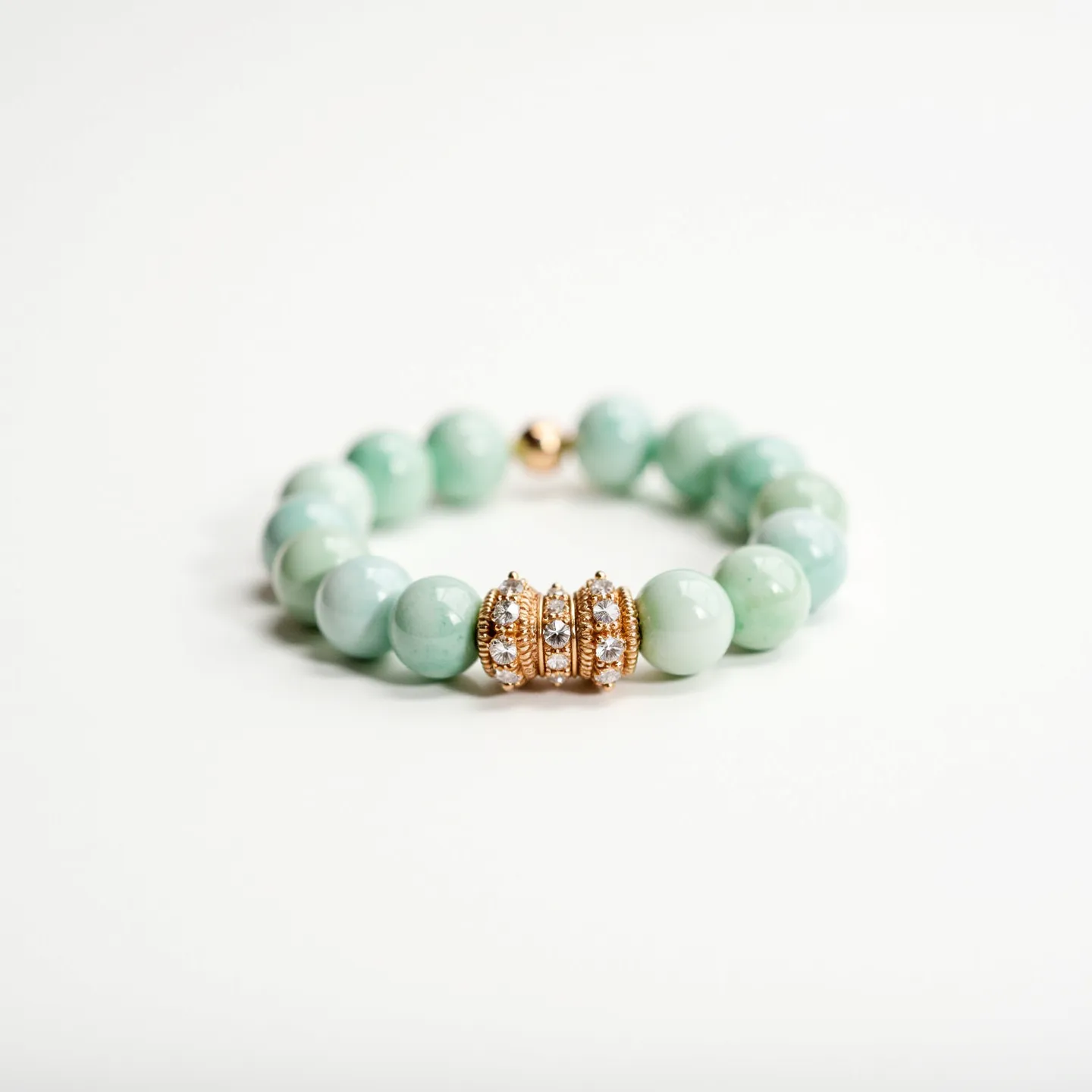 This baby bracelet features polished, round, light green beads, likely made of a semi-precious stone such as jade or aventurine. The centerpiece is a trio of gold-toned metal spacers adorned with small, clear gemstones set in a pavé style, providing a sparkling accent. The bracelet appears to be held together with an elastic band, ensuring a flexible and comfortable fit for a baby, without a traditional clasp mechanism. The combination of smooth stones and glittering gems gives the piece a charming and elegant look suitable for a young child.