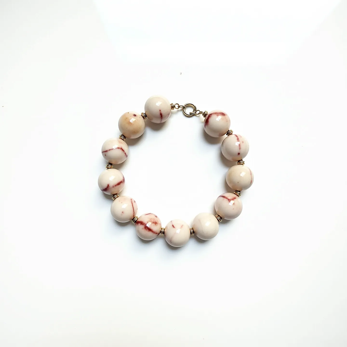 This baby bracelet features a series of round, polished beads that appear to be made of a creamy white material with red streaks or marbling, giving them a subtle and natural look. The beads are strung together with small gold-colored spacer rings that add a touch of elegance between each bead. The bracelet is secured with a simple yet functional gold-colored metal clasp, likely a lobster clasp, which ensures it stays securely fastened while being easy to open and close. The overall design is delicate and playful, suitable for a baby.