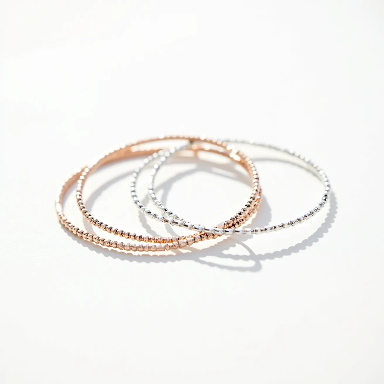 This bangles feature a delicate design comprising two rose gold toned bangles and one white toned bangle, each formed from a series of small, evenly spaced, spherical elements. The bangles appear to be made of metal, exhibiting a subtle shine indicative of a polished surface. There are no visible gems or stones embedded in the bangles, and the surface texture is consistent throughout. The bangles are a continuous loop with no visible clasps or attachments, suggesting a slip-on style suitable for easy wear.