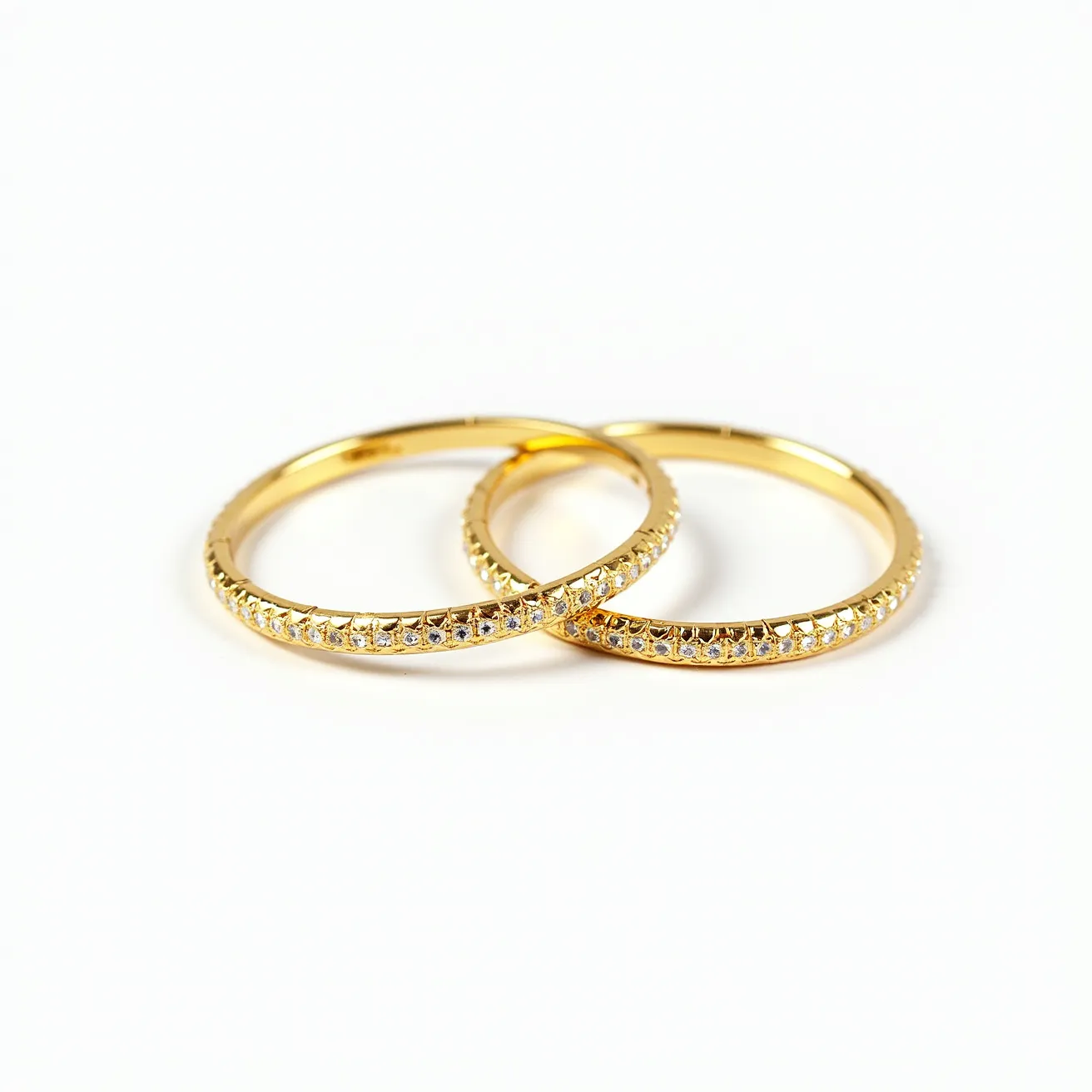 These bangles are crafted from a gold-colored metal, adorned with a row of small, clear, round-cut gemstones set in a continuous pattern along the circumference. The stones are set in a prong setting, which secures them in place while allowing maximum light to pass through for added sparkle. The bangles appear to be smooth on the inner side for comfortable wear and do not feature any clasps or visible attachments, suggesting a slip-on design. The overall style is elegant and would complement a variety of outfits.