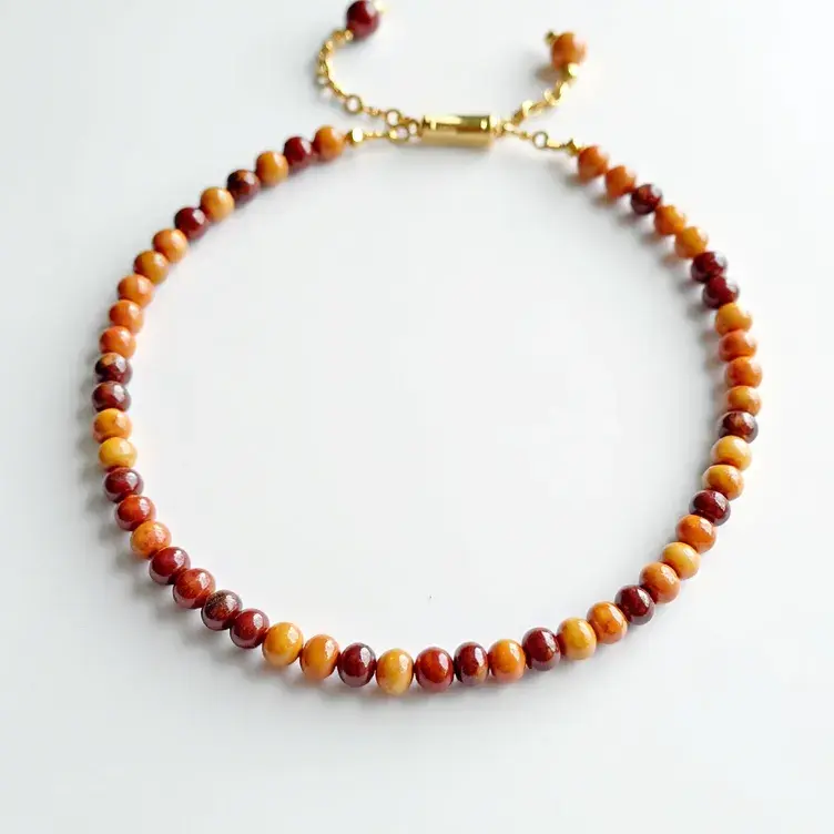 This beaded bracelet features a series of polished beads in shades of red, yellow, and brown. The beads are spherical and uniform in size, likely crafted from dyed or natural stones. The bracelet is secured with a gold-toned lobster clasp that attaches to an adjustable chain, allowing for a customizable fit. The chain ends are adorned with small beads that match the primary ones, adding a cohesive and decorative touch. The overall design is simple yet elegant, making it a versatile accessory for various outfits and occasions.