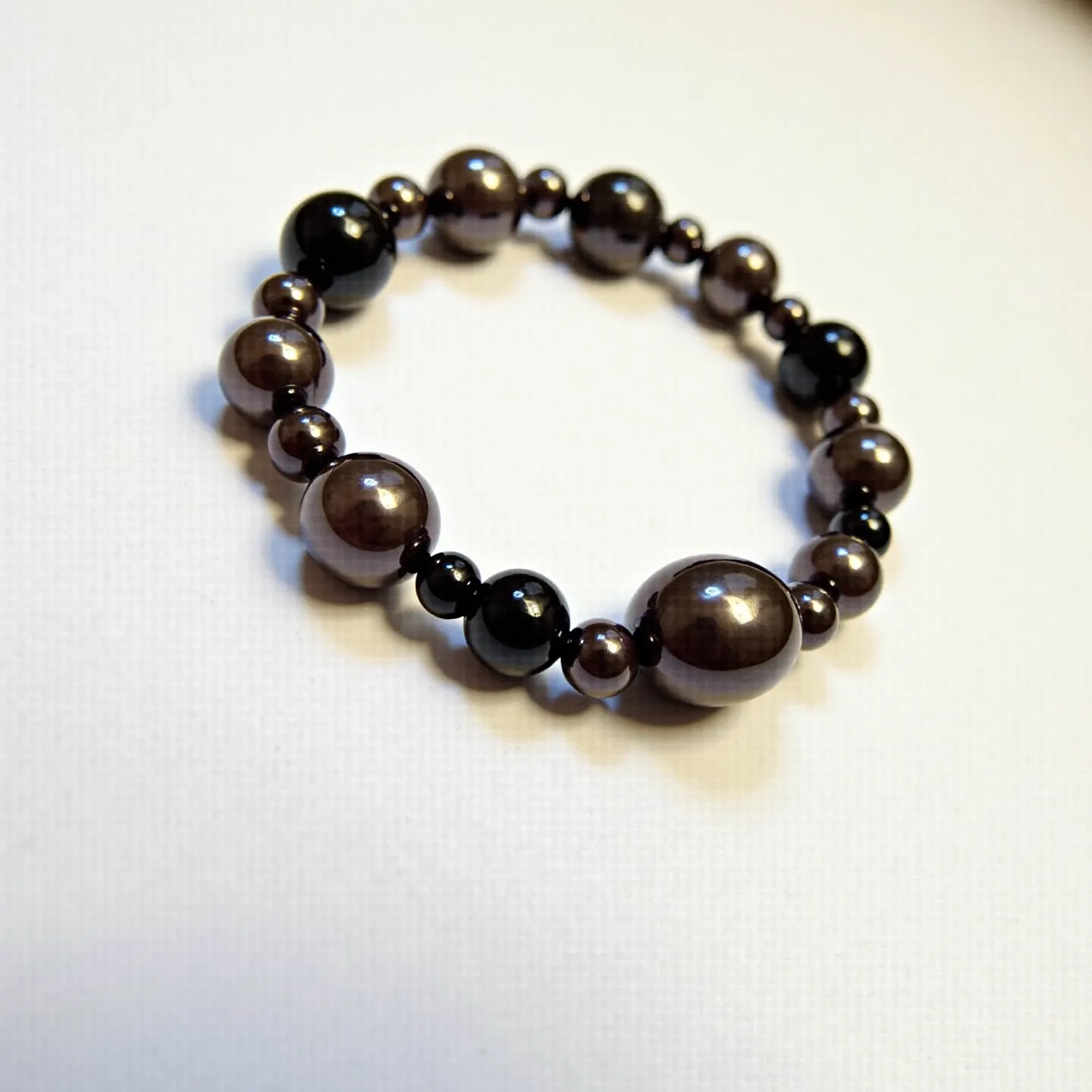 This beaded bracelet features a sequence of round, polished beads in varying sizes, which appear to be made from materials with a lustrous finish, possibly hematite or glass. The beads are dark in color, with some showing a metallic sheen and others a more subdued matte finish. They are strung together on an elastic cord, allowing for a stretchy fit without the need for a clasp or specific attachment mechanism. The overall design is simple yet elegant, making it a versatile accessory for various occasions.