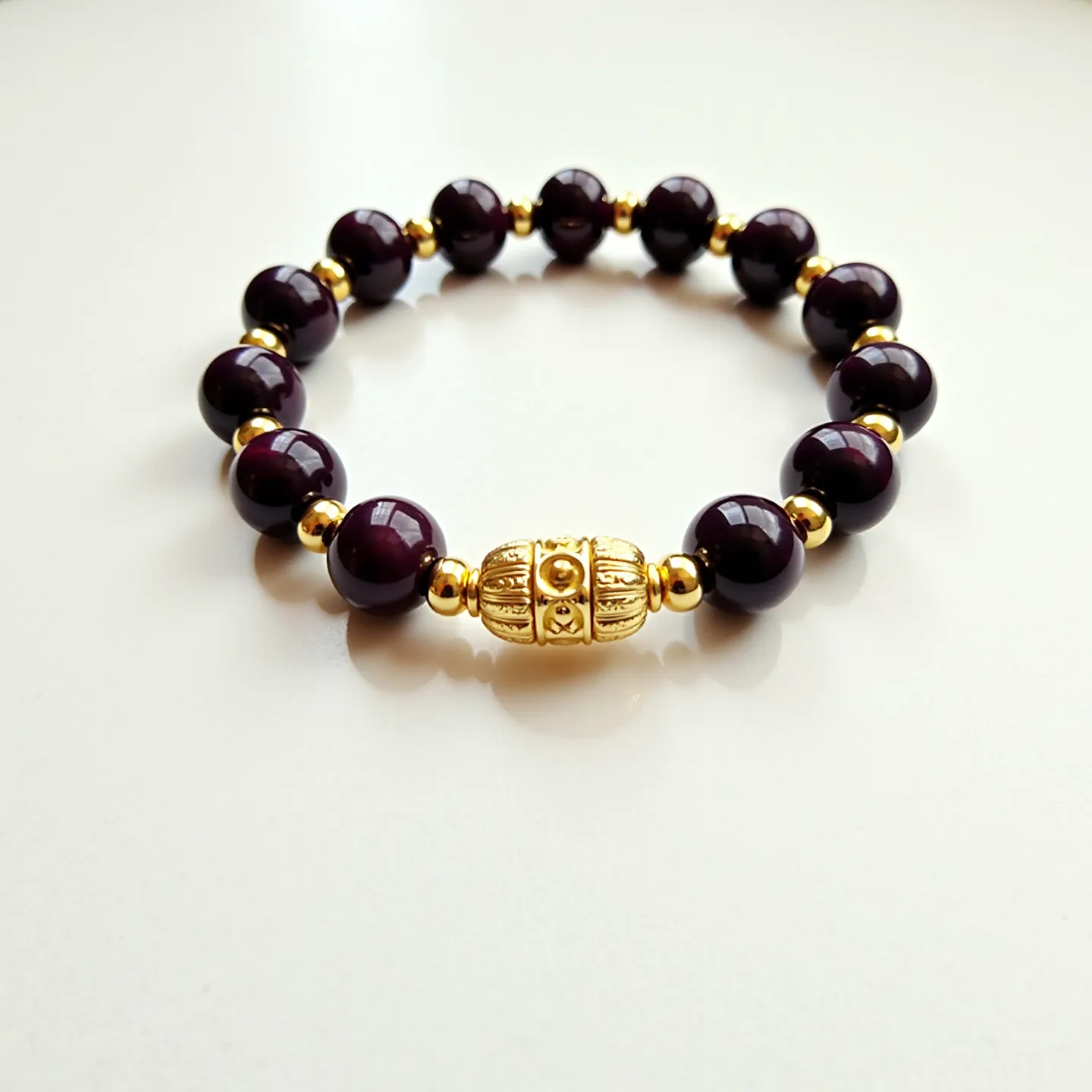 This beaded bracelet features a series of smooth, dark-colored beads that appear to be made of a polished stone, strung together with small gold-colored spacer beads interspersed between them. The dark beads are symmetrical and spherical, contributing to the bracelet’s uniform appearance. The central piece is a larger, intricately designed gold-colored bead with ornamental patterns, serving as both a focal point and a statement piece. The bracelet does not feature a traditional clasp, suggesting it might be elastic or stretchable for easy wear.