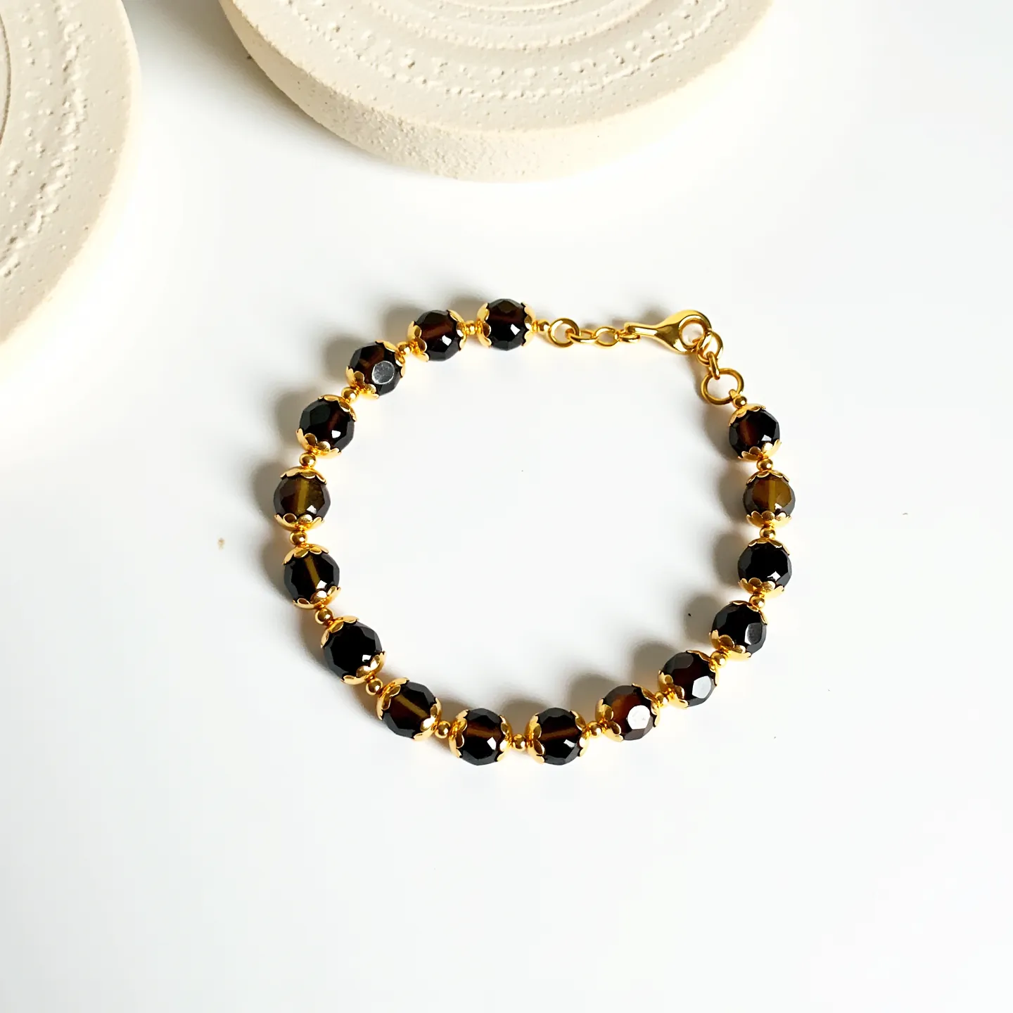 This beaded bracelet features intricately faceted dark beads that could be various types of gemstone or glass, each separated by small golden spacer beads. The beads are uniformly shaped and exhibit a high-gloss finish, enhancing their polished appearance. The bracelet is assembled using golden wire which integrates seamlessly with its elegant clasp, a lobster claw, providing a secure and functional closure. The meticulous construction reflects careful craftsmanship, resulting in a refined and stylish piece of jewelry.