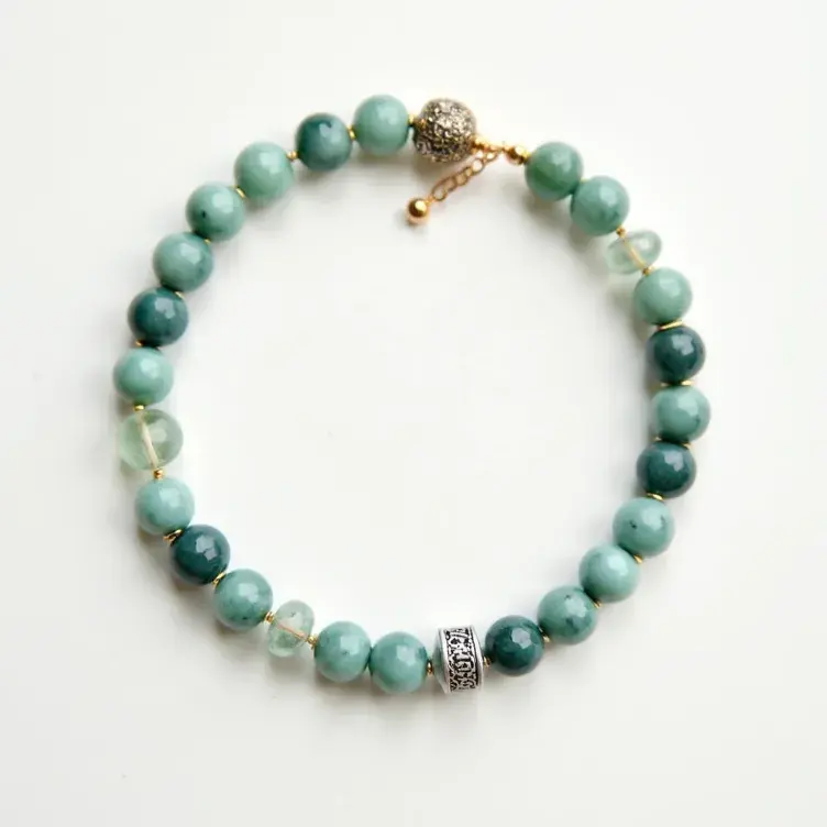 This beaded bracelet features an arrangement of uniformly sized, polished, round beads in varying shades of light to dark green, interspersed with clear, faceted beads for accentuation. The beads appear to be made of a semi-precious stone, possibly jade or turquoise, complemented by small gold spacer beads. A single, intricately designed silver bead acts as a centerpiece, adding to the overall aesthetic appeal. The bracelet is secured with a gold-tone chain and clasp, which provides an adjustable and secure fit. A decorative filigree round bead near the clasp adds an elegant finishing touch.