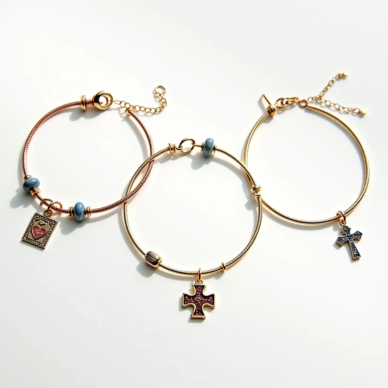 This charm bracelet set features three distinct styles, each adorned with unique charms and intricate details. The first bracelet is a rose gold tone bangle with a rectangular charm that has a detailed heart motif, bordered by silver-toned elements and flanked by two blue ceramic or glass beads. The second bracelet is a yellow gold tone bangle with a charm shaped in the form of a cross, encrusted with small red stones in a pavé setting. The third bracelet also exhibits a yellow gold tone but showcases a simpler design with a single cross charm embellished with blue stones, again in a pavé setting. Each bracelet is equipped with a different clasp mechanism: the first exhibits a lobster clasp with an adjustable chain, the second has a simplistic hook clasp, and the third also uses a lobster clasp with an adjustable chain for secure fastening.