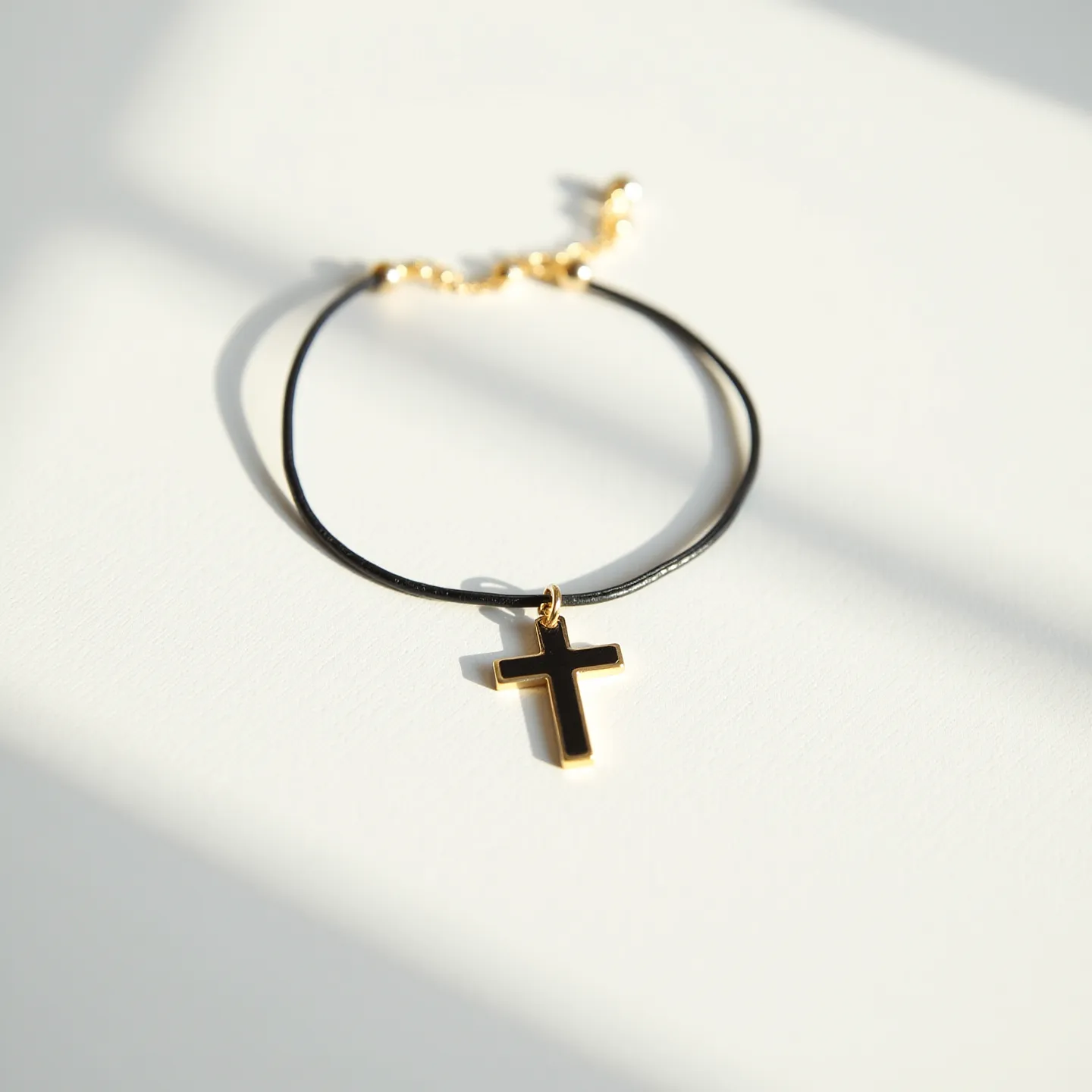 This cross bracelet features a black cross pendant outlined in gold, suspended from a slender, black band. The band appears to be made of a smooth, leather-like material and is attached to a gold link chain that allows for adjustable sizing, secured with a small round gold clasp. The overall aesthetic is a combination of simplicity and elegance, enhanced by the interplay of the black and gold tones.