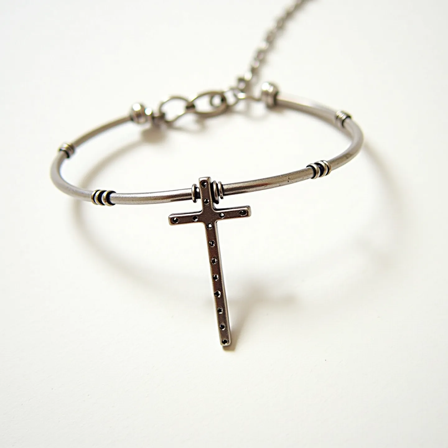 This cross bracelet features a sleek and minimalist design crafted from a metallic material, likely stainless steel. The bracelet sports a thin, circular band with small decorative bands that add to its aesthetic value. Dangling from the middle of the band is a simple cross adorned with evenly spaced small round accents, which might be tiny stud-like elements rather than gems. The cross is attached to the band via a small hoop, allowing it to move freely. The bracelet is secured with a lobster clasp that connects to an adjustable chain, ensuring a snug and custom fit for the wearer.