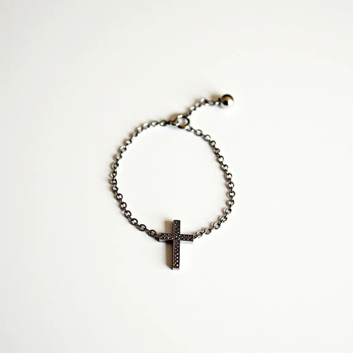 This cross bracelet features a sleek design crafted from a metal chain and showcases a cross pendant at its center. The cross is intricately adorned with small, sparkling gemstones set in a paved arrangement, enhancing its visual appeal. The bracelet is equipped with a simple sliding ball clasp that allows for easy adjustment of the fit, combining both aesthetic charm and practicality.