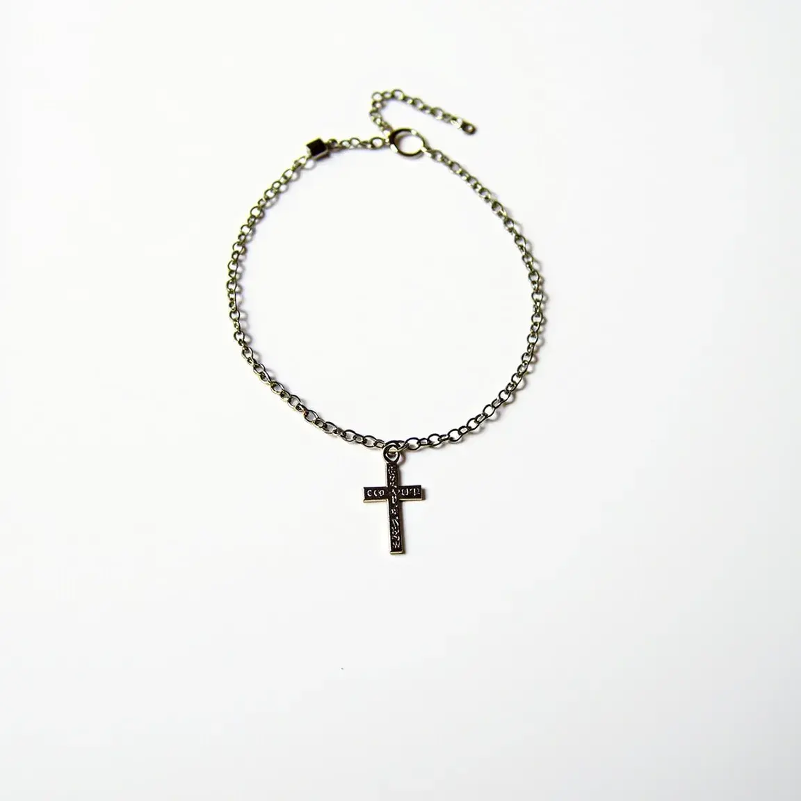 This cross bracelet features a delicate chain made of a metallic material, possibly stainless steel or another alloy, which links together in a series of small, rounded links. The bracelet includes a cross-shaped pendant that hangs centrally from a link in the chain. This pendant appears to be made from the same metal as the chain and is rectangular in shape, featuring inscriptions or engravings along the horizontal and vertical bars of the cross. The bracelet is secured with a lobster clasp fastening, accompanied by an adjustable chain section with additional links to allow for size customization.