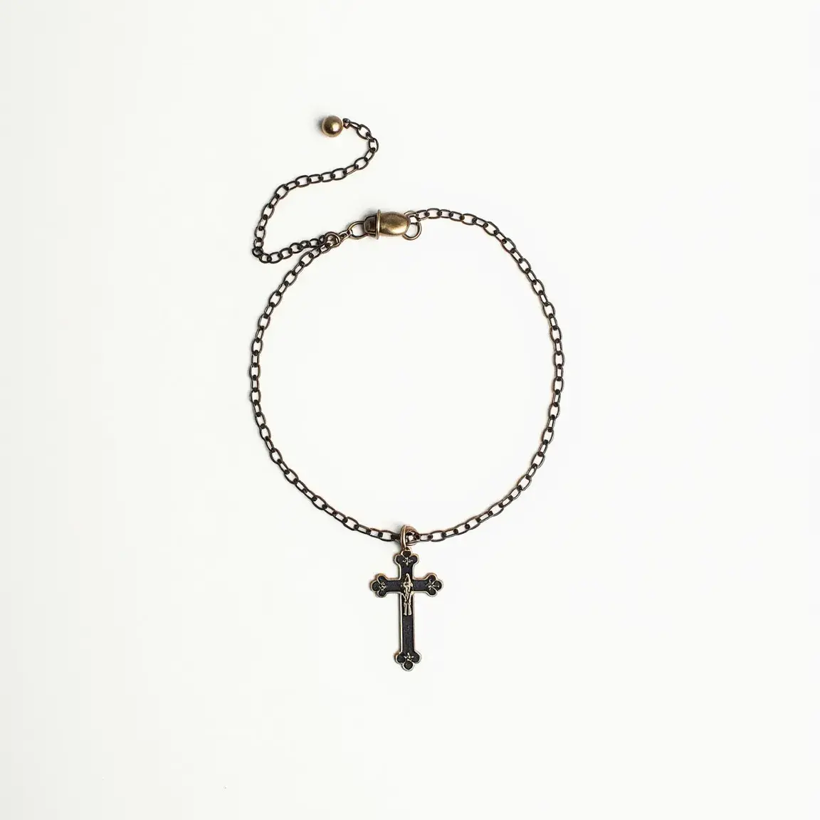 This cross bracelet features a delicate metal chain, most likely made of brass or bronze, with a hanging cross pendant. The cross is detailed with small, rounded tips and appears to be inset with dark, possibly black, stones that add a subtle shine. The bracelet is equipped with an adjustable closure, which includes a chain with a small metal ball at the end, allowing for a custom fit on the wrist. The simple yet elegant design of the cross and the fine detailing of the chain make it a striking piece.