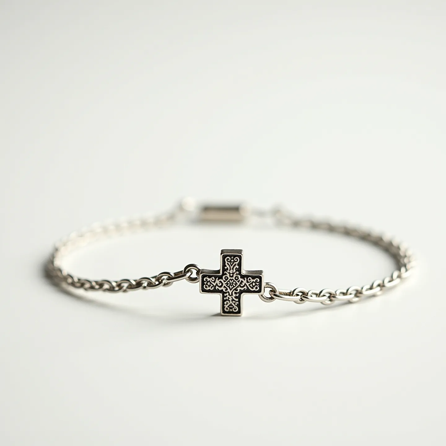 This cross bracelet features a finely detailed metal cross centerpiece with intricate engraved patterns, which hangs from a slender, polished chain. The bracelet is likely made from silver or a silver-toned metal, giving it a sleek and sophisticated appearance. It is secured with a cylindrical clasp that completes the minimalist yet elegant design. The absence of gemstones or additional embellishments keeps the focus on the detailed craftsmanship of the cross.