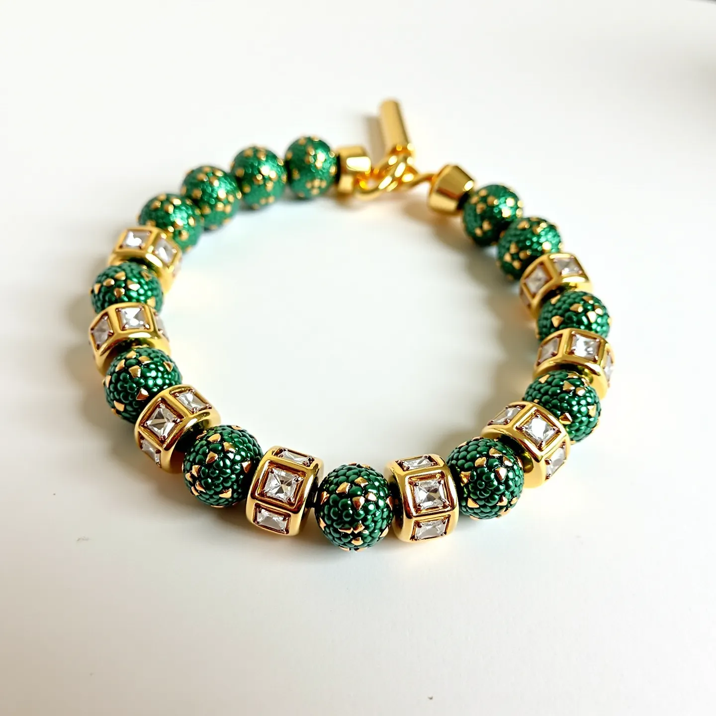 This gold beaded bracelet features intricately designed green beaded spheres, interspersed with square-cut clear gems set into gold. The gems are housed in gold settings that provide a stark contrast and add elegance to the design. The bracelet is secured with a gold clasp that ensures a firm and stylish closure, making it a striking piece of jewelry that combines vibrant color and luxurious materials.