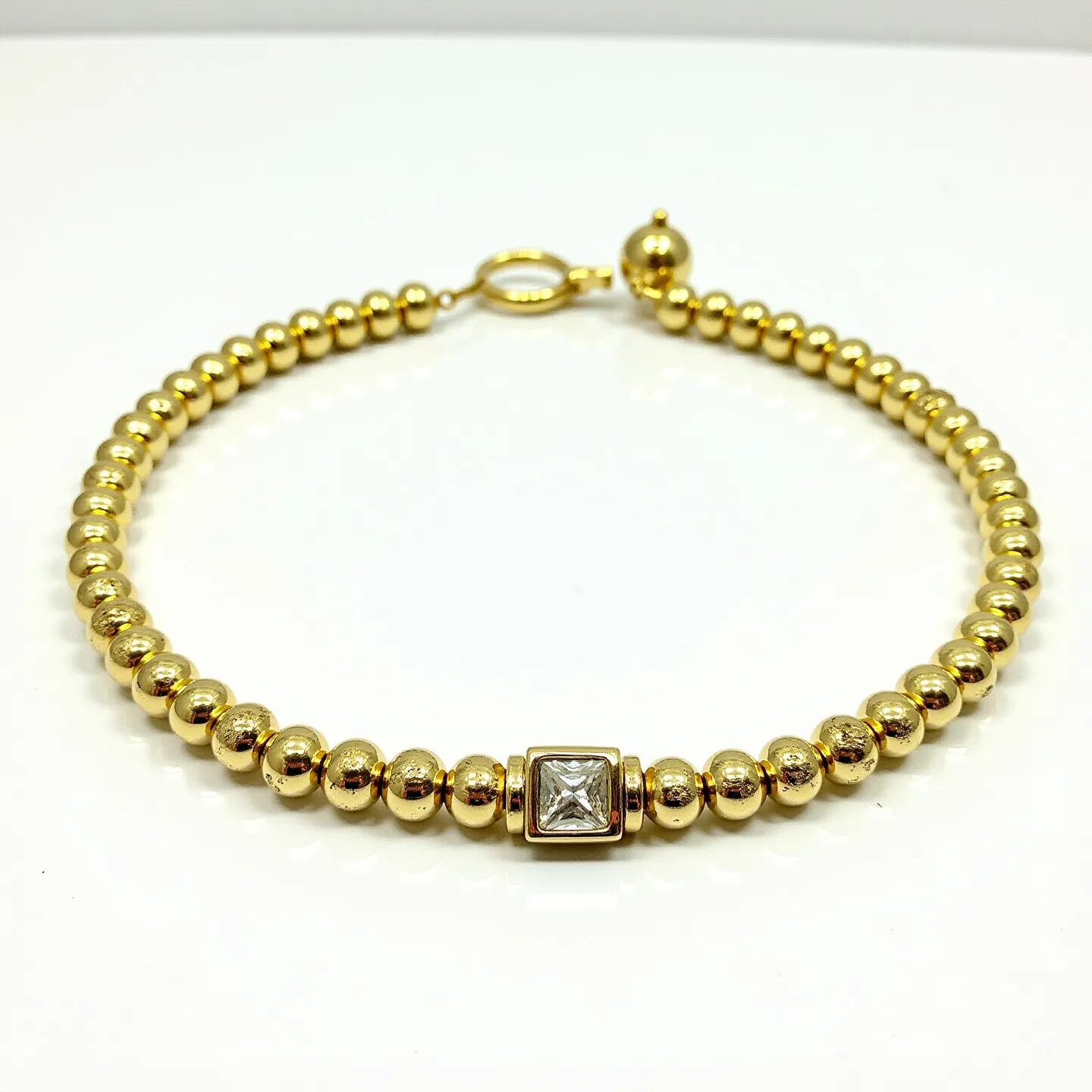 This gold beaded bracelet features a series of polished spherical beads crafted from gold, creating a continuous loop of glistening orbs. At its centerpiece, a square setting houses a single, faceted gemstone, cut in a brilliant square shape. The stone is securely held in place by a bezel setting, complementing the overall design with a touch of sparkle. The bracelet fastens with a gold ring clasp, ensuring a secure and elegant closure.