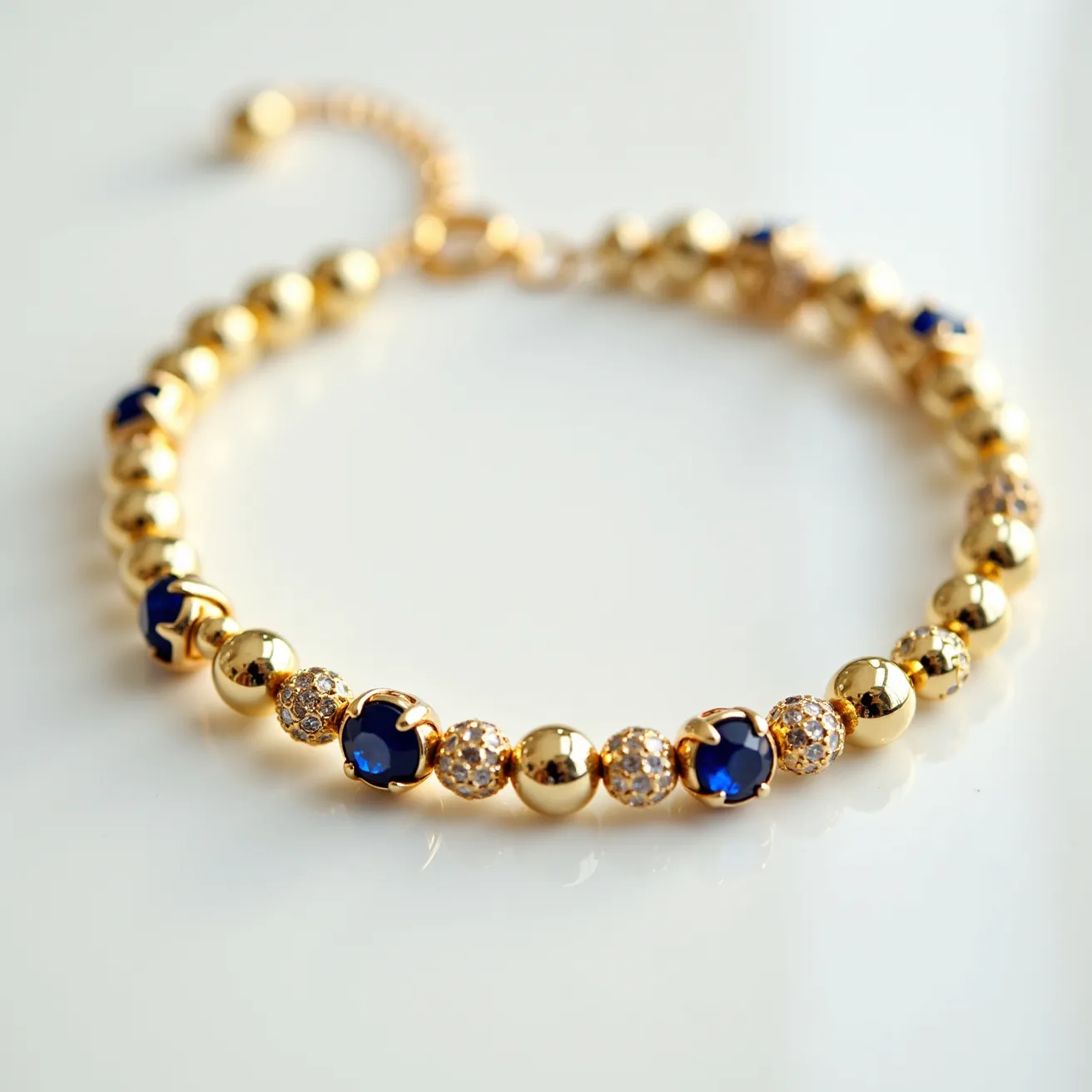 This gold beaded bracelet features a series of uniformly-sized, polished gold beads interspersed with larger, blue gemstones that appear to be sapphires. The sapphires are round-cut and held in a prong setting, which allows the stones to catch light from various angles. Additionally, the bracelet incorporates small, clear stones, likely diamonds, set in a pave style to add sparkle and contrast. The bracelet is equipped with an adjustable chain clasp, which allows for a secure and customized fit. The combination of gold, blue sapphires, and clear stones gives this bracelet an elegant and luxurious appearance.