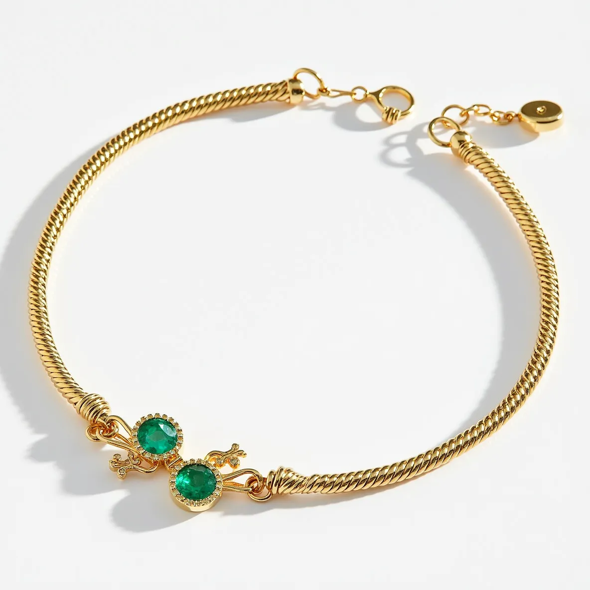 This gold beaded bracelet features a twisted rope design made of gold, showcasing intricate craftsmanship. At its center are two round-cut green gemstones set in bezel settings, which are likely emeralds given their distinct color. The clasp is a simple yet secure spring ring type, ensuring easy wear and removal. A small, circular charm adds an extra element of elegance, completing the design with a touch of sophistication.