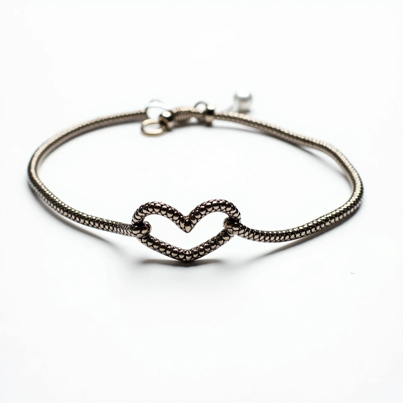 This heart bracelet features a textured metal band that forms a continuous loop, ending in a heart-shaped centerpiece. The heart and the band both showcase an intricate pattern of small, raised motifs, giving the piece an elegant and refined appearance. The bracelet is fastened with a lobster clasp, providing a secure closure. The design is minimalist yet sophisticated, making it a versatile accessory for various occasions.