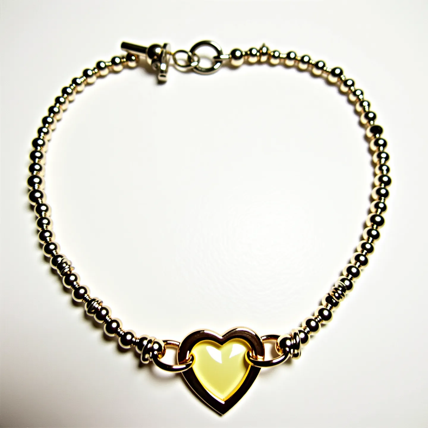 This heart bracelet features polished metallic beads strung together to form the main band, providing a sleek and reflective surface. The centerpiece of the bracelet is a heart-shaped charm showcasing a smooth, glossy yellow stone, which is meticulously cut and set within a metallic frame. The heart charm is securely connected to the bracelet with finely crafted loops. The bracelet is fastened with a toggle clasp, ensuring both ease of use and a secure fit when worn.