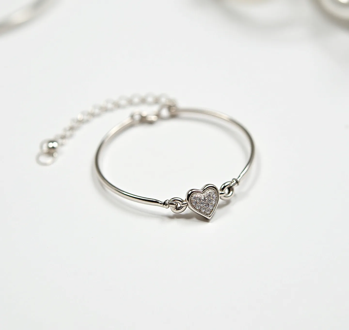 This heart bracelet features a delicate metal band, likely made of sterling silver or white gold, designed in a minimalist circular shape. At its center, it showcases a heart-shaped charm adorned with multiple small, clear gemstones set in a pavé style, which provides a sparkling, uniform look across the heart surface. The attachment method includes a fine chain extension with a simple round clasp, suggesting adjustability in the bracelet's length for a comfortable fit. The heart charm is securely connected to the band via small loops on either side, ensuring stability and elegance in its design.