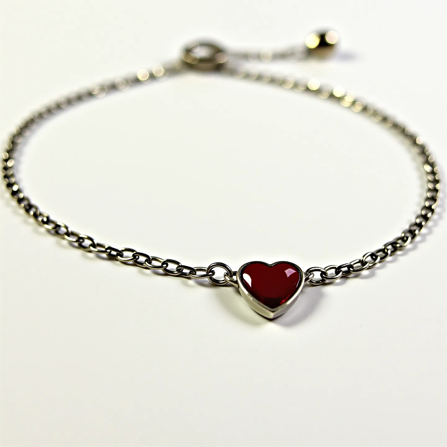 This heart bracelet features a delicate chain made of a metallic material, likely silver or stainless steel, characterized by small, evenly spaced links. At the center of the bracelet is a heart-shaped centerpiece with a smooth, polished red gemstone or enamel set in a bezel setting, which provides a secure and defined border around the stone. The bracelet is equipped with an adjustable slider clasp, allowing for a customizable fit, and the clasp is complemented by small, rounded metal charms for added detail. The overall design is elegant and minimalistic, with the red heart element serving as a striking focal point.