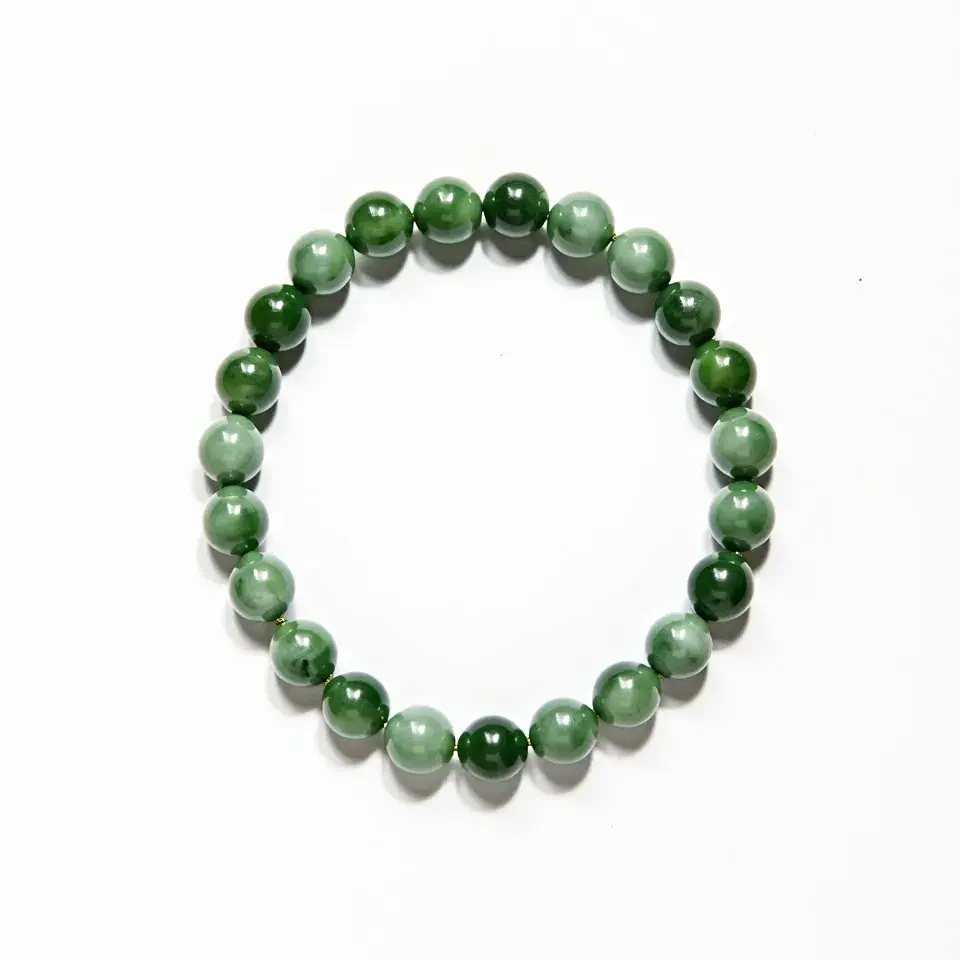 This jade bracelet features a series of polished round jade beads, each exhibiting a smooth and glossy finish. The beads are uniformly sized and display varying shades of green, characteristic of natural jade. There are no additional gems, stones, or decorative settings present. The bracelet is held together by an elastic string, allowing it to stretch and fit comfortably around the wrist without the need for a clasp or other attachment mechanism.