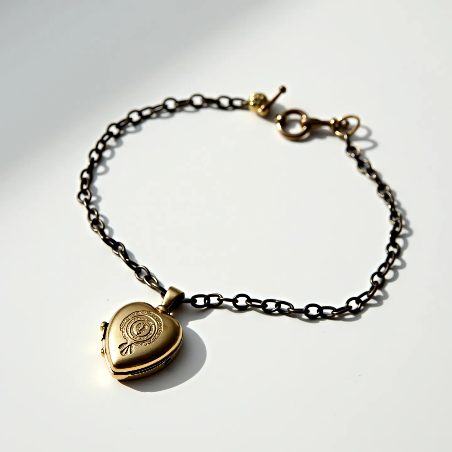 This locket bracelet features a delicate chain made of blackened metal links, lending it a vintage and elegant appearance. The centerpiece is a heart-shaped locket crafted from a gold-toned metal, which appears to be engraved with a subtle floral design. The locket is affixed to the chain by a small matching gold-toned loop. The bracelet secures with a toggle clasp, incorporating both a circular ring and a bar, also in a complementary gold-toned finish, providing both functionality and aesthetic coherence. The craftsmanship and material choice contribute to its overall charm and appeal.