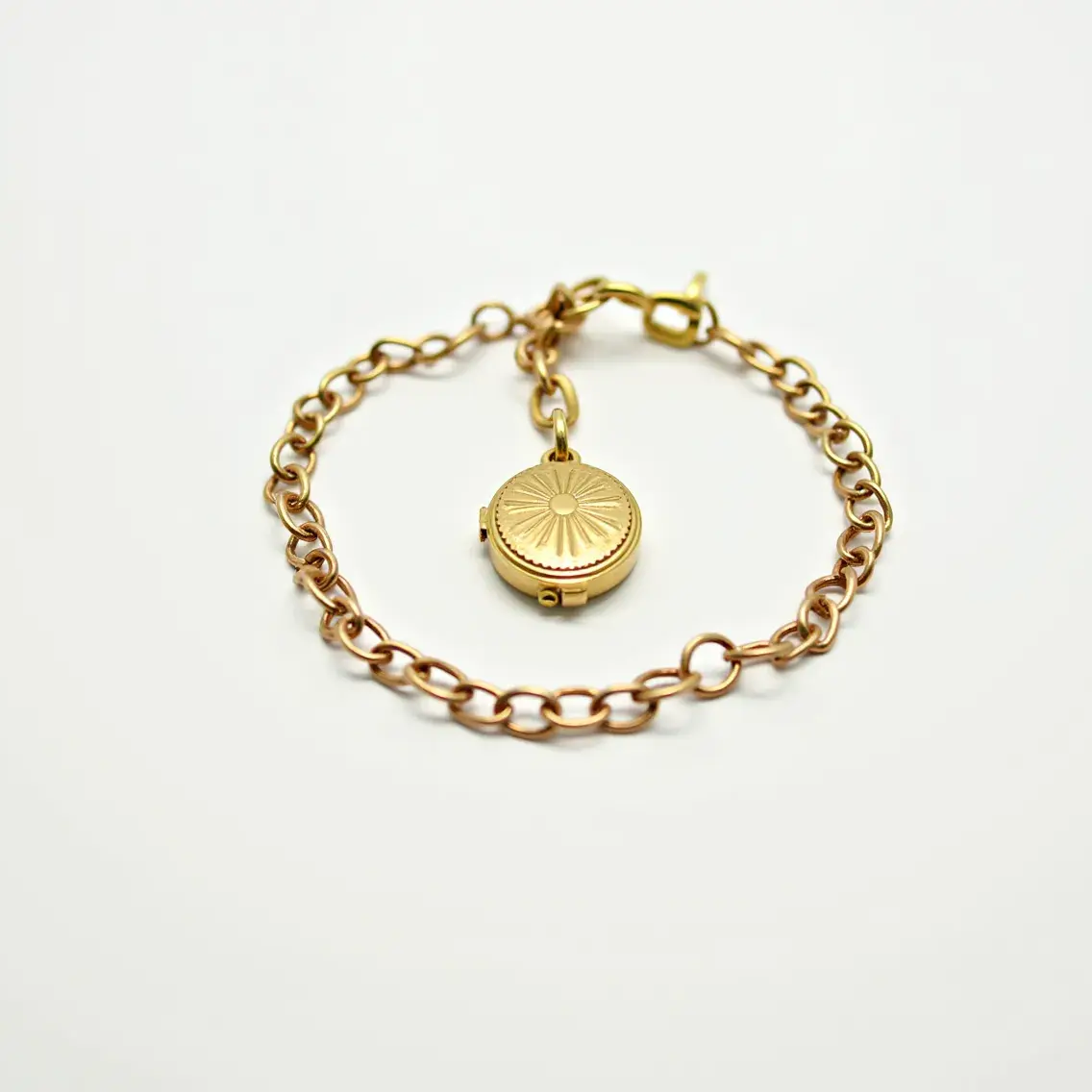 This locket bracelet features a delicate chain made of what appears to be gold, displaying a warm, yellow-toned metallic finish. At its focal point hangs a small, round locket adorned with an intricate radial sunburst pattern on its surface, adding an elegant decorative touch. The locket is securely attached to the chain with a matching gold loop. The bracelet is closed with a lobster clasp, ensuring a secure and adjustable fit on the wrist. The overall design is both classic and timeless, highlighting the beauty and subtle charm typical of vintage-inspired jewelry pieces.