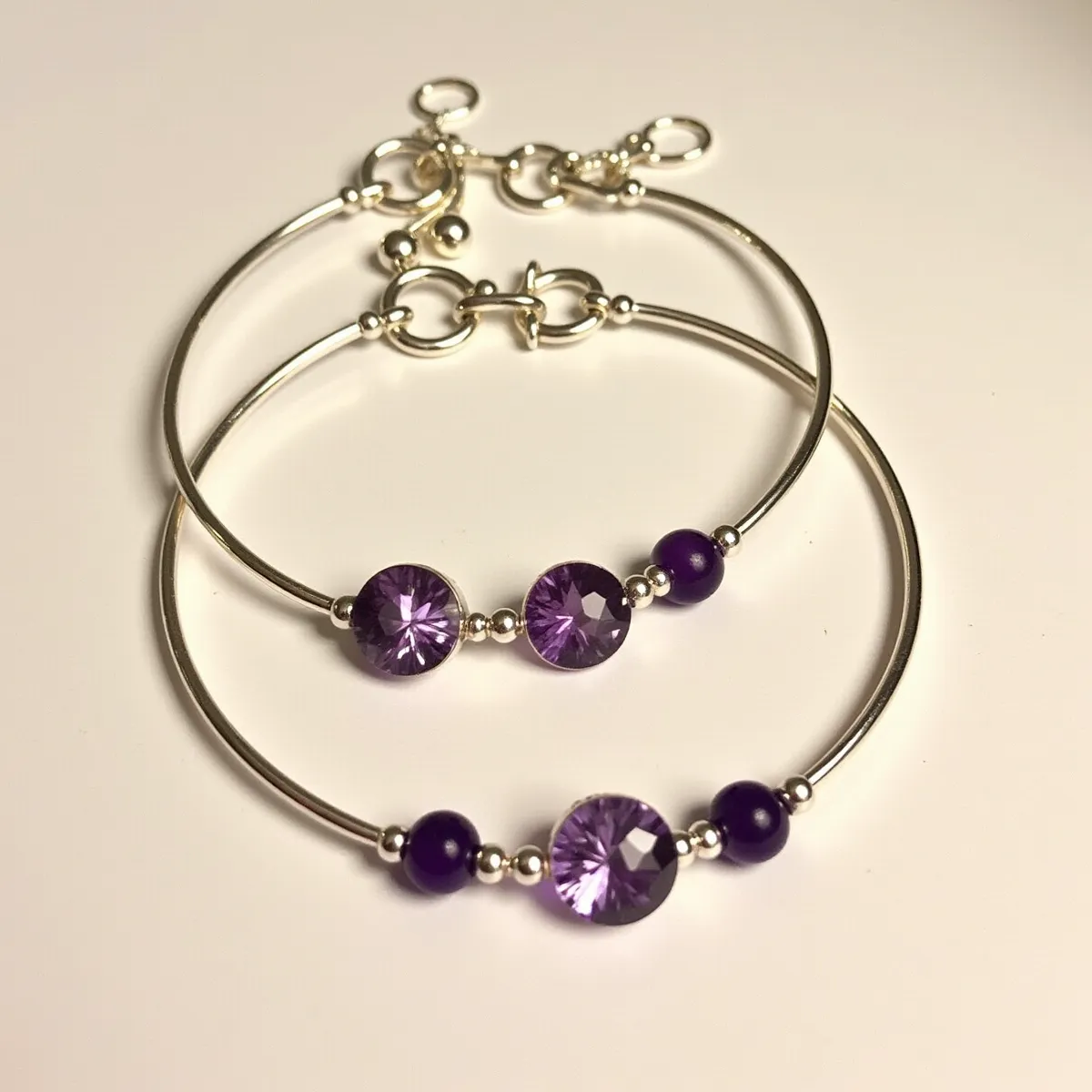 This matching bracelets feature a sleek and simple silver bangle design. Each bracelet is adorned with a round cut purple gemstone, set in a bezel setting, which showcases its faceted brilliance. Flanking the central gemstone are smaller round beads in a deep purple hue, adding a complementary accent to the larger gem. The bracelets are secured with a toggle clasp, featuring circular links that align with the overall elegant theme of the jewelry. The materials used in these bracelets create a cohesive and stylish appearance suitable for various occasions.