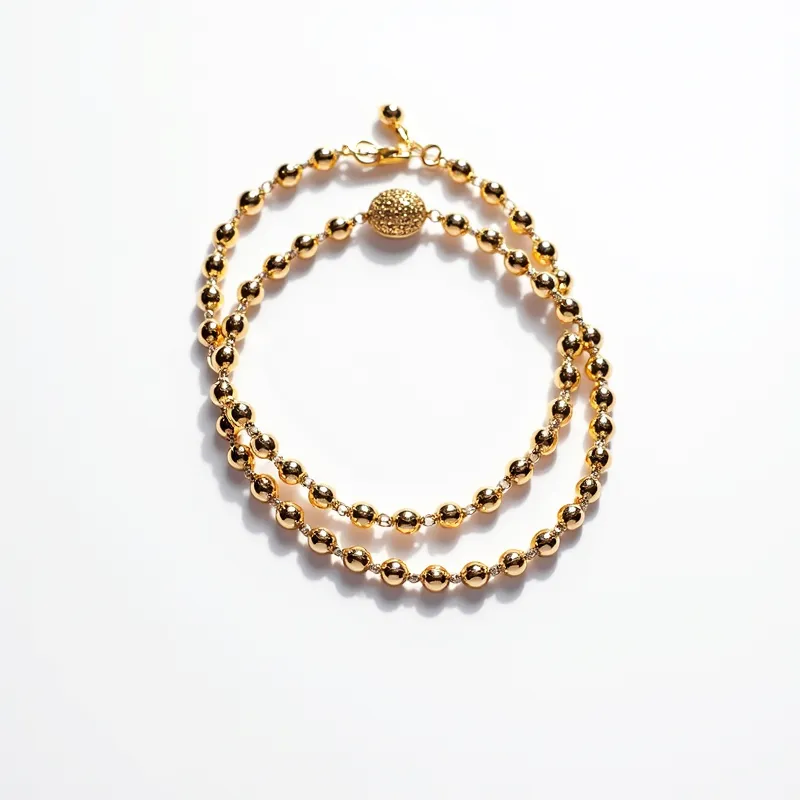 This matching bracelets are composed of shiny gold-toned beads strung together in a simple yet elegant design. Each bracelet features uniformly shaped, spherical beads that reflect light beautifully. Centrally, there is a larger, textured bead that stands out with a more intricate surface compared to the rest of the smooth beads, adding a touch of visual interest. The bracelets are secured with a decorative gold-toned ball clasp that not only ensures they stay in place but also complements their overall aesthetic. The combination of polished and textured elements makes these bracelets appear refined and stylish.