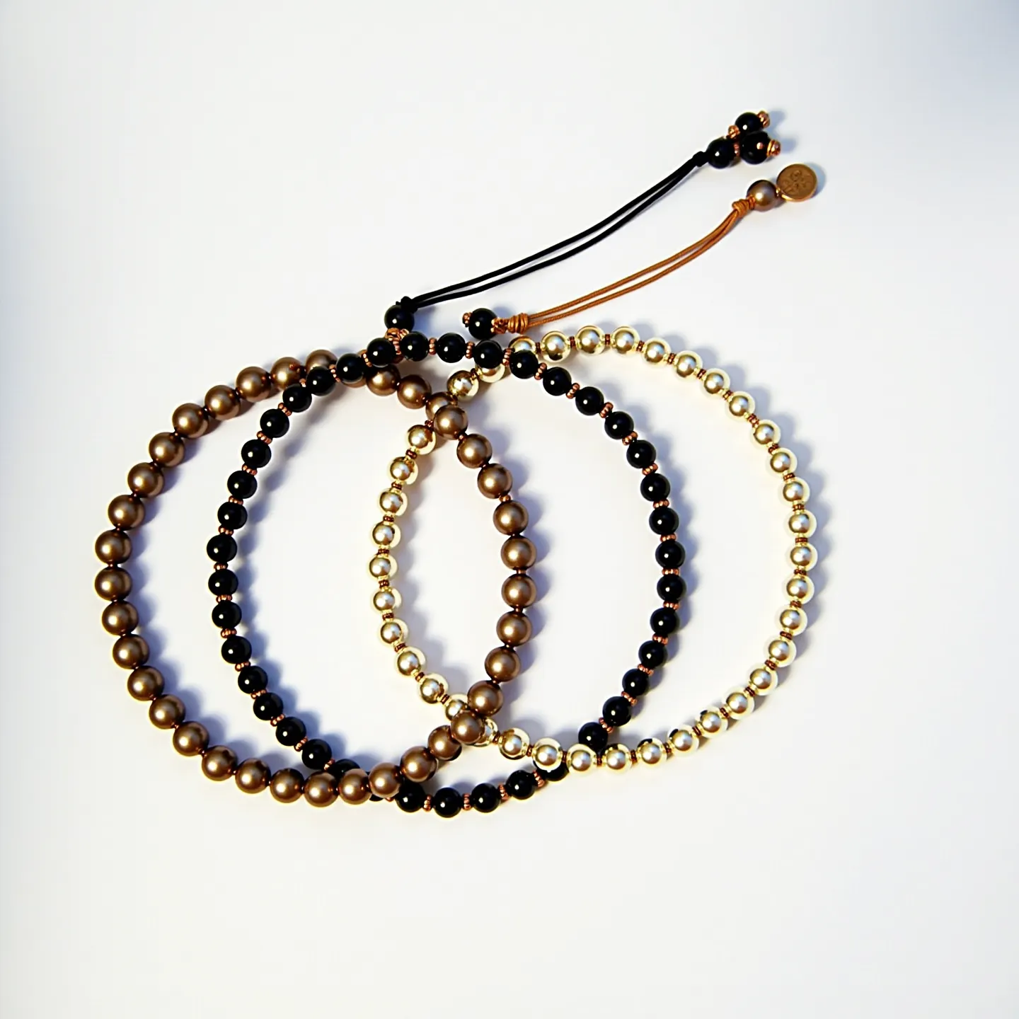 This matching bracelets showcase an elegant design, featuring round beads in three distinct color themes: a bronze tone, a gold tone, and black beads. Each bracelet is composed of uniformly sized beads strung together, with alternating patterns of black and respective colored beads. The bracelets are secured with a knot-and-loop closure mechanism, utilizing black and brown cords. Additionally, decorative beads and a small disc are integrated into the closure for a finishing touch. The beads themselves appear polished, contributing to an overall sophisticated and coordinated aesthetic.