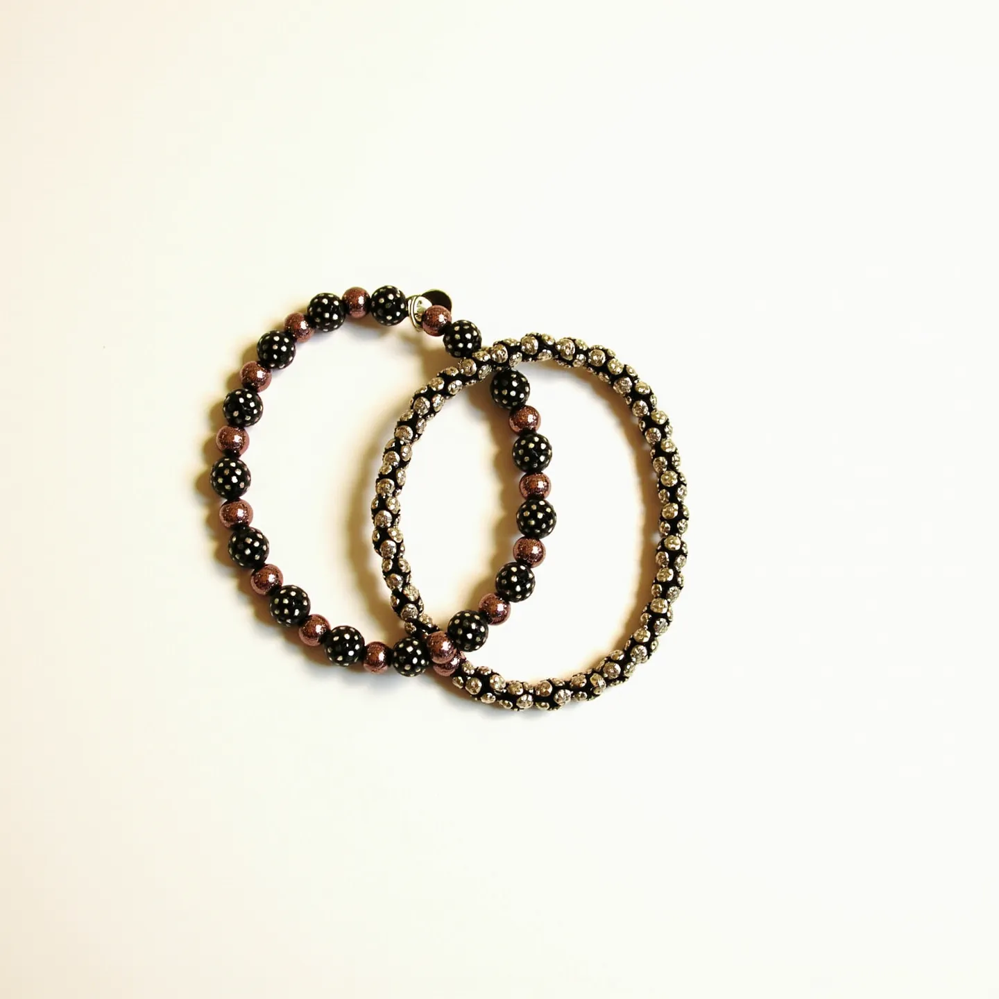 This matching bracelets feature an arrangement of alternating black beads with white polka dots and metallic-like brown beads. Each black bead is uniformly adorned with numerous small white dots, giving a playful yet sophisticated look. The bracelets are designed with an elastic band, allowing them to easily stretch over the wrist for a comfortable fit. The secondary bracelet incorporates the same black and brown beads but is distinguished by a sparkling accent of small, clear, round-cut crystals set in a continuous chain that encircles the entire length of the bracelet, adding an element of elegance. The construction appears sturdy with the gems and beads securely set and strung together.