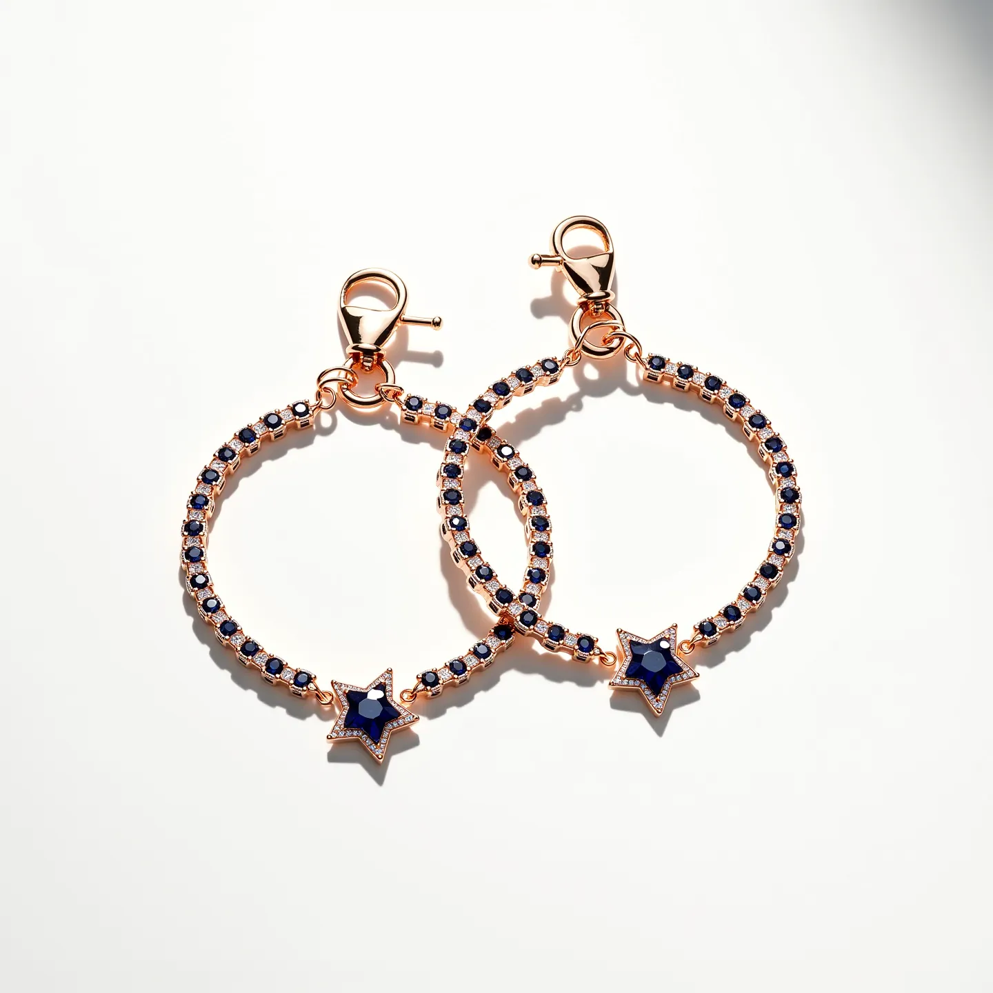 This matching bracelets feature a circular design embellished with numerous small, round-cut blue gems set into a base material that appears to be a rose-gold metal. Each bracelet is adorned with a star-shaped charm, also encrusted with similar blue stones, adding a focal point to the piece. The bracelets are designed with a lobster clasp, providing a secure and easy fastening option. The combination of the blue gems and rose-gold setting gives the bracelets an elegant and stylish appearance.