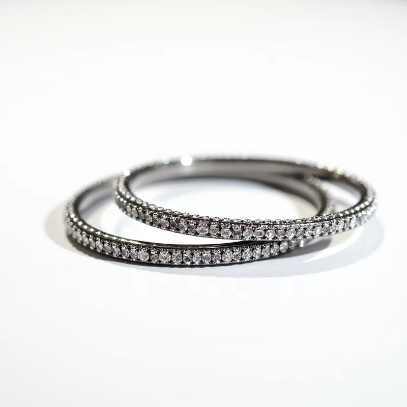 This matching bracelets are intricately crafted from a polished metal, likely silver or white gold, and adorned with a continuous line of small, round-cut gems set into the design. The gems, which appear to be diamonds or diamond-like stones, are uniformly set in a prong setting, enhancing their brilliance and allowing maximum light reflection. The bracelets have a sleek, circular band design without any noticeable clasp, suggesting they are slip-on bangles. The uniformity and precision of the gem arrangement add an elegant sparkle to the overall design, making them suitable for both sophisticated and casual wear.