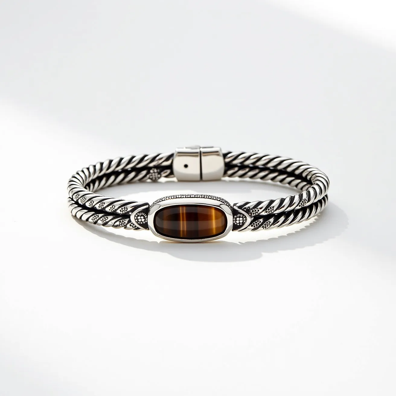 This men's bracelet features a twisted metal rope design, predominantly composed of silver-toned material. The central element is an oval gemstone, likely a tiger's eye, showcasing rich, brownish hues with natural striping. The stone is cabochon cut and securely set in a polished bezel frame. The bracelet includes intricate detailing around the stone setting, with patterned embellishments extending along the band. It secures with a robust, streamlined clasp mechanism, ensuring both functionality and aesthetic cohesion.