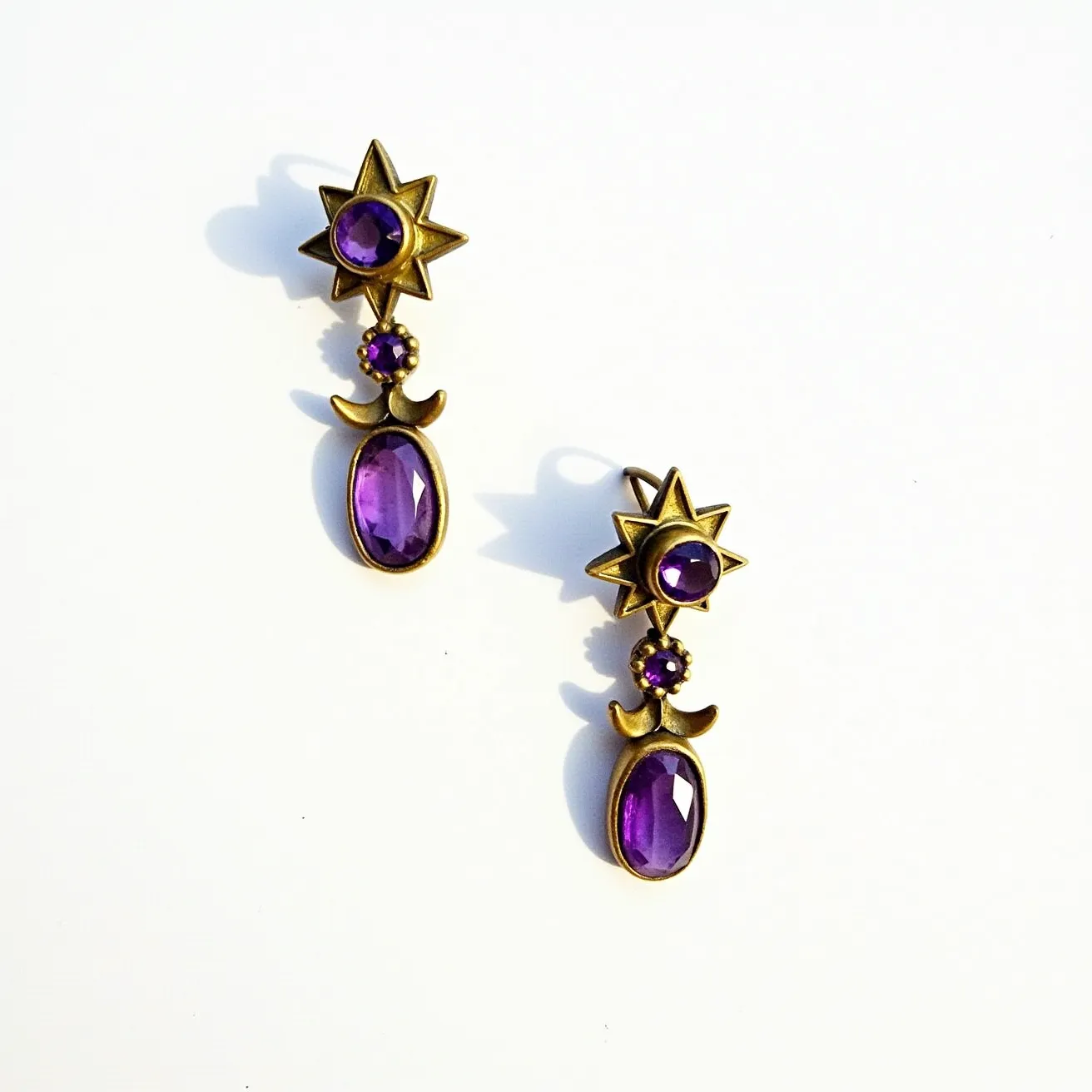 These 70s earrings feature a striking design with a star motif made from a metallic material, likely aged brass, which gives them a vintage look. Each earring incorporates three purple stones: a round-cut stone in the center of the star, a small round-cut stone connected beneath it, and an oval-cut stone as the prominent drop. The stones are likely set in bezels that match the overall metallic tone, enhancing the cohesive aesthetic. The clasp or attachment seems to be a classic post-back style, commonly used for pierced ears during that era.