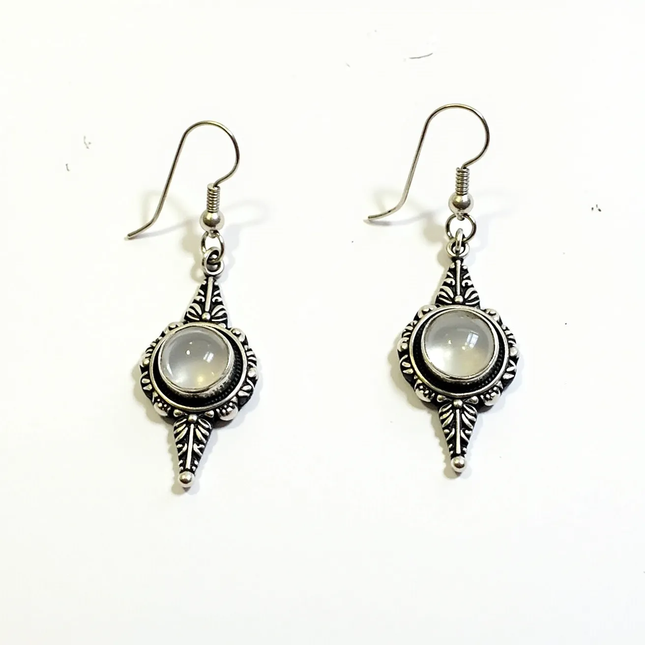 These 70s earrings features intricate metalwork likely made from a silver-toned material, showcasing a bohemian style typical of the era. At the center of each earring, a cabochon-cut gem with a smooth, rounded surface adds a subtle elegance, suggesting the use of a moonstone or similar gem. The stone is set within a decorative bezel that complements the ornate design around it, with symmetrical leaf-like patterns and small bead details enhancing the vintage aesthetic. These earrings use a fishhook clasp, allowing for easy attachment and a comfortable fit.