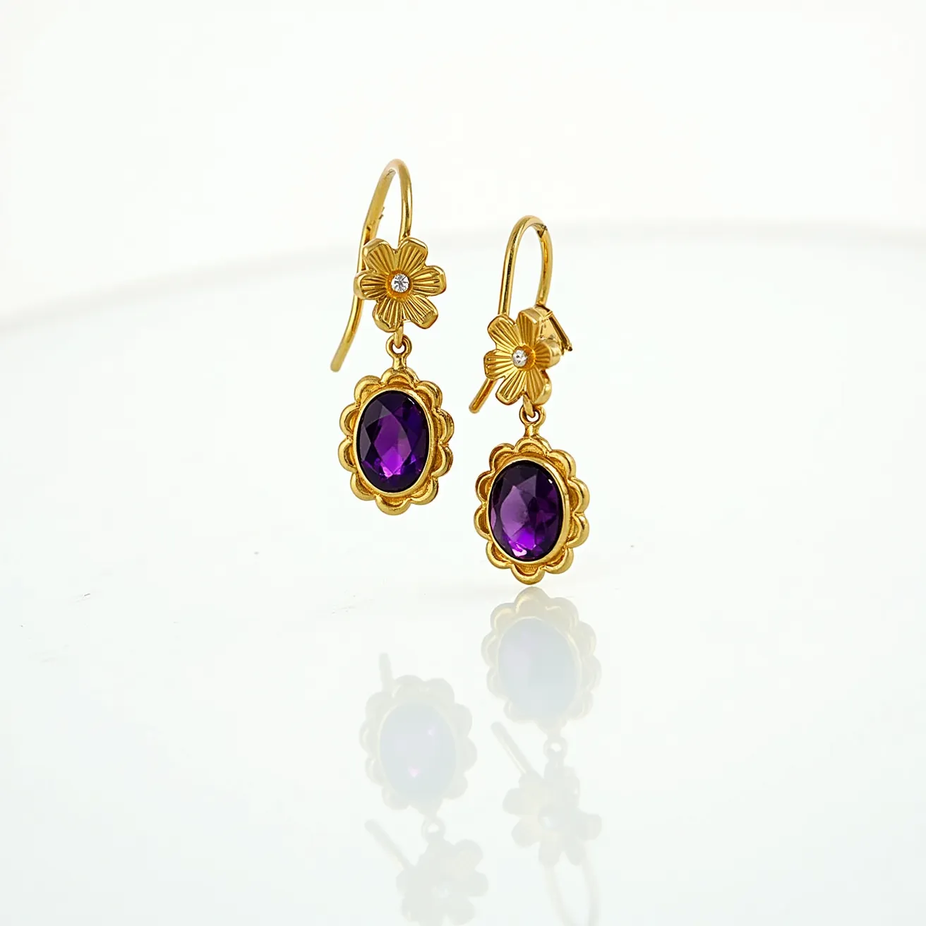 These 70s earrings feature a striking design characterized by vibrant, oval-cut amethyst-like gemstones nestled in a scalloped gold-toned setting. The gemstones are faceted, allowing them to catch the light beautifully. Above the gemstones, each earring is adorned with a floral element, featuring petals that highlight a central, clear, round-cut stone, likely a small diamond or diamond substitute. The earrings are suspended from a fish hook clasp, ensuring a secure yet elegant attachment, making them both stylish and functional. The combination of the deep purple gem and the warm tones of the metal creates a classic look reminiscent of the era's bold aesthetic.