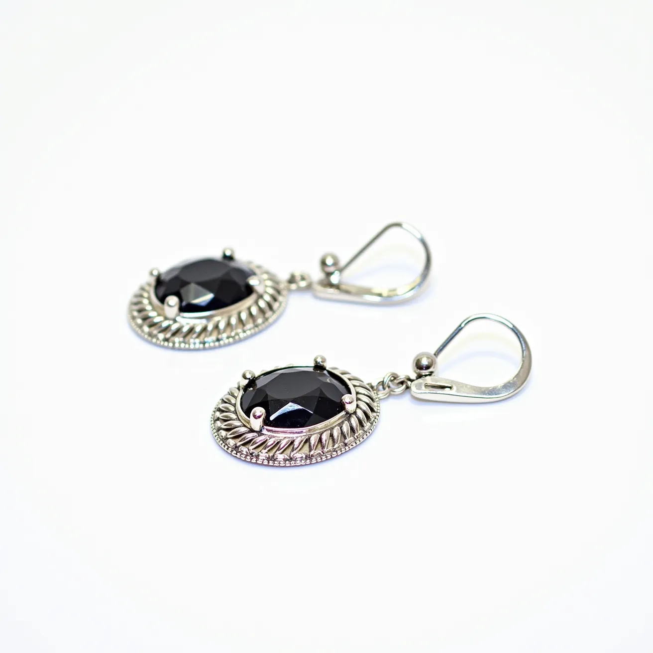 These 70s earrings feature a striking design with large black gemstones, possibly onyx or a dark crystal, set in an oval shape. The stones are faceted, which adds a touch of sophistication, and are secured with a prong setting. The surrounding metalwork exhibits intricate rope-like detailing, enhancing the earring's vintage appeal. Crafted from what appears to be a silver-toned metal, these earrings are both bold and elegant. They are equipped with lever-back clasps, ensuring a secure and comfortable fit for the wearer.