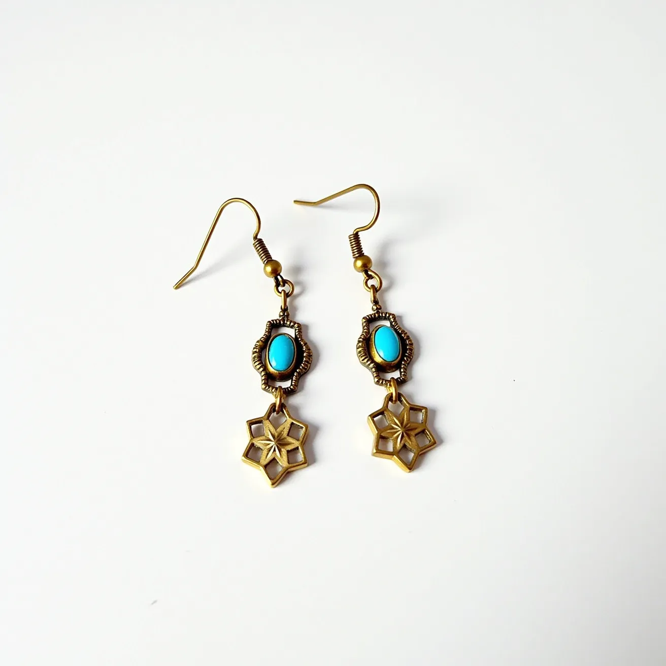 These 70s earrings design features a notable combination of materials and craftsmanship, incorporating a bright turquoise-colored stone centrally set in an oval shape. The stone appears to be set within an ornate brass or gold-toned metal frame, characterized by intricate detailing that enhances its vintage appeal. Dangling below the stone is a geometric star-shaped charm, which complements the overall aesthetic, adding a touch of whimsy and elegance. The earrings use a simple fishhook clasp for attachment, a popular choice for its ease of wear and suitability for dangle earring styles of the era. The harmonious blend of turquoise and metal gives these earrings a distinctive look that resonates with the bold, expressive fashion trends of the 1970s.