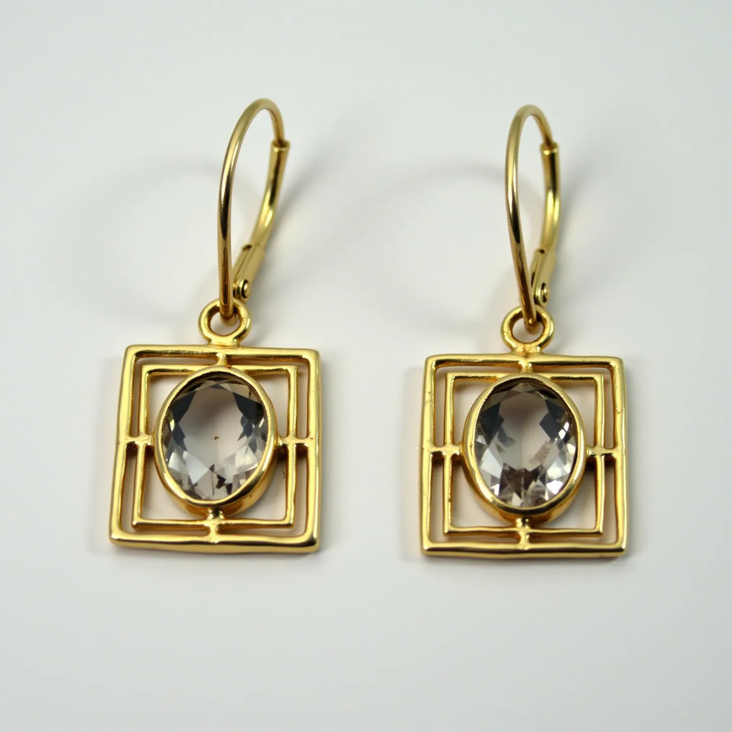 These 70s earrings feature a striking geometric design with gold-toned metal forming a double rectangular frame. At the center of each earring is an oval-shaped faceted gem, set in a bezel, which elegantly captures light. The stones appear to be clear, resembling quartz or possibly a similar crystal, adding a subtle sparkle to the overall look. These earrings are finished with a lever-back clasp for secure attachment, aligning with the retro style of the era. The design highlights a blend of minimalism and boldness, characteristic of 1970s jewelry aesthetics.