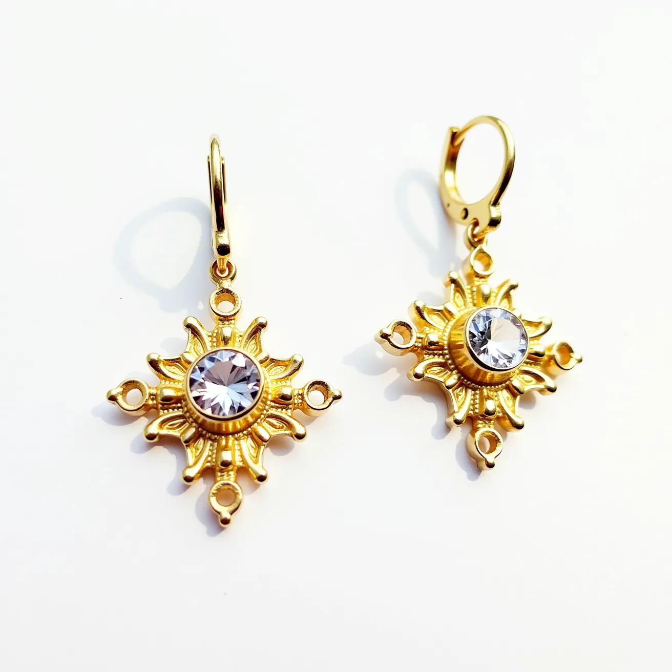 These 70s earrings showcase an intricate sunburst design crafted in gold-toned metal. At the center of each earring, a round, faceted crystal gem is bezel-set, catching the light with its clear, sparkling surface. The elaborate detailing around the gem features radiating beams with small loops at the ends, adding to the decorative elegance. These earrings are equipped with lever-back clasps, ensuring secure attachment while enhancing the overall vintage style. The combination of the warm metallic tones with the brilliance of the crystal evokes a classic, glamorous appeal typical of 1970s jewelry fashion.